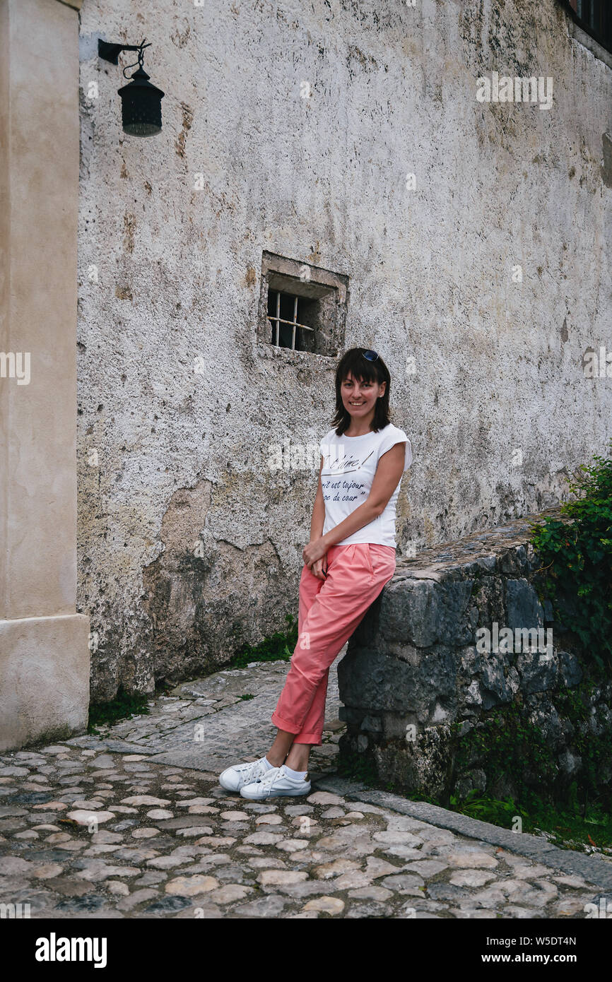Junge lächelnde Frau Touristen sitzen auf einer Burg von Bled Wand im Innenhof. Tragen weiße Hemd, weiße Turnschuhe und Koralle Hose. Travel Concept Stockfoto