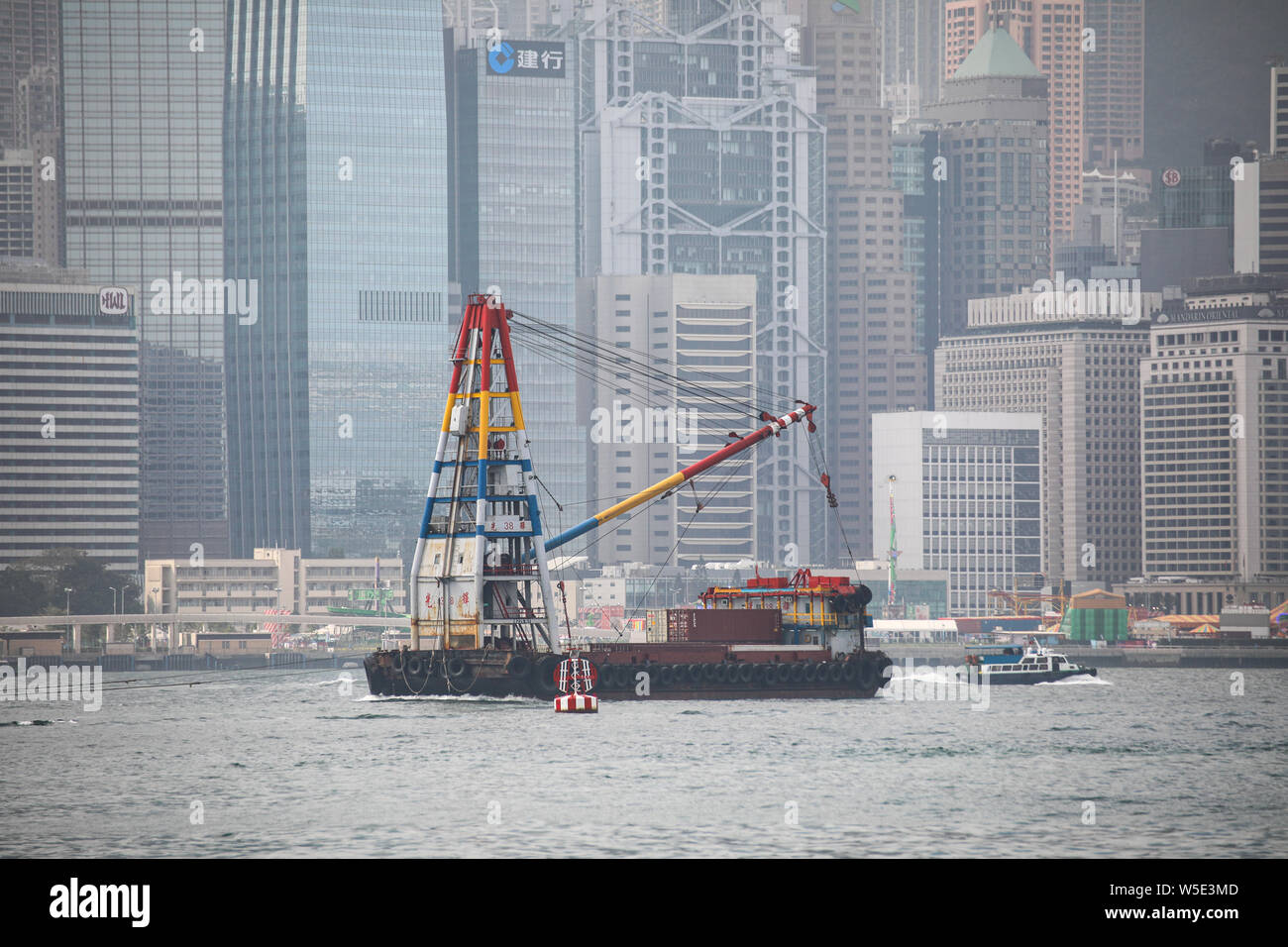 Container Barge mit Derrick Kran im Victoria Harbour und Hong Kong Island Wolkenkratzer auf Hintergrund Stockfoto