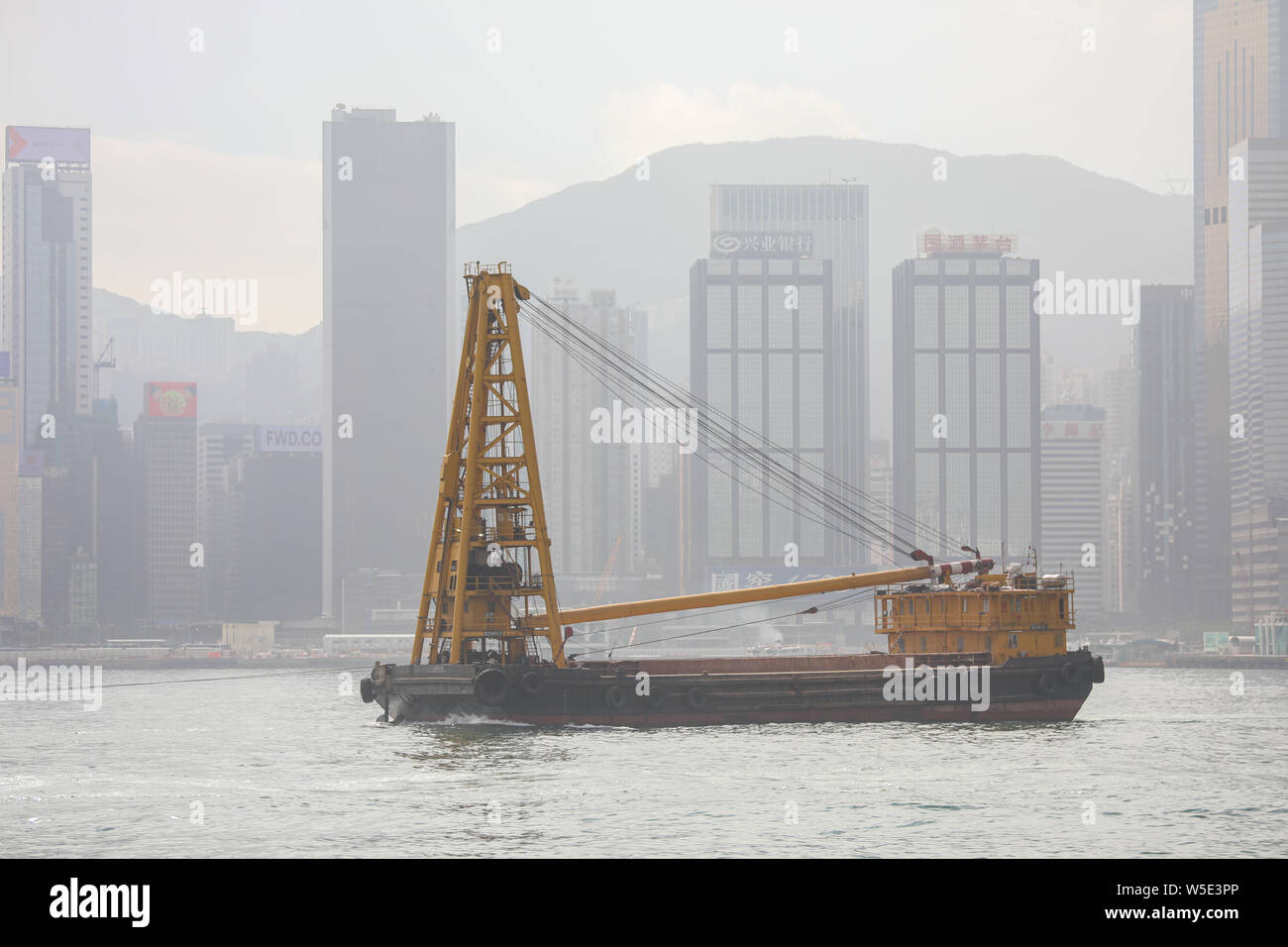 Container Barge mit Derrick Kran auf Victoria Harbour und Hong Kong Island auf Hintergrund Stockfoto
