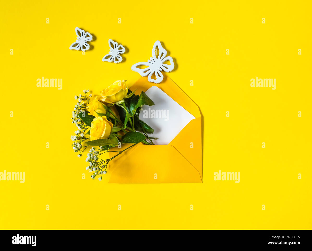 Gelbe Rosen und weißen Gypsophila in Umschlag und drei Holz- Schmetterlinge close-up auf gelben Hintergrund. Ansicht von oben, flach. Vorlage für eine Begrüßung Stockfoto