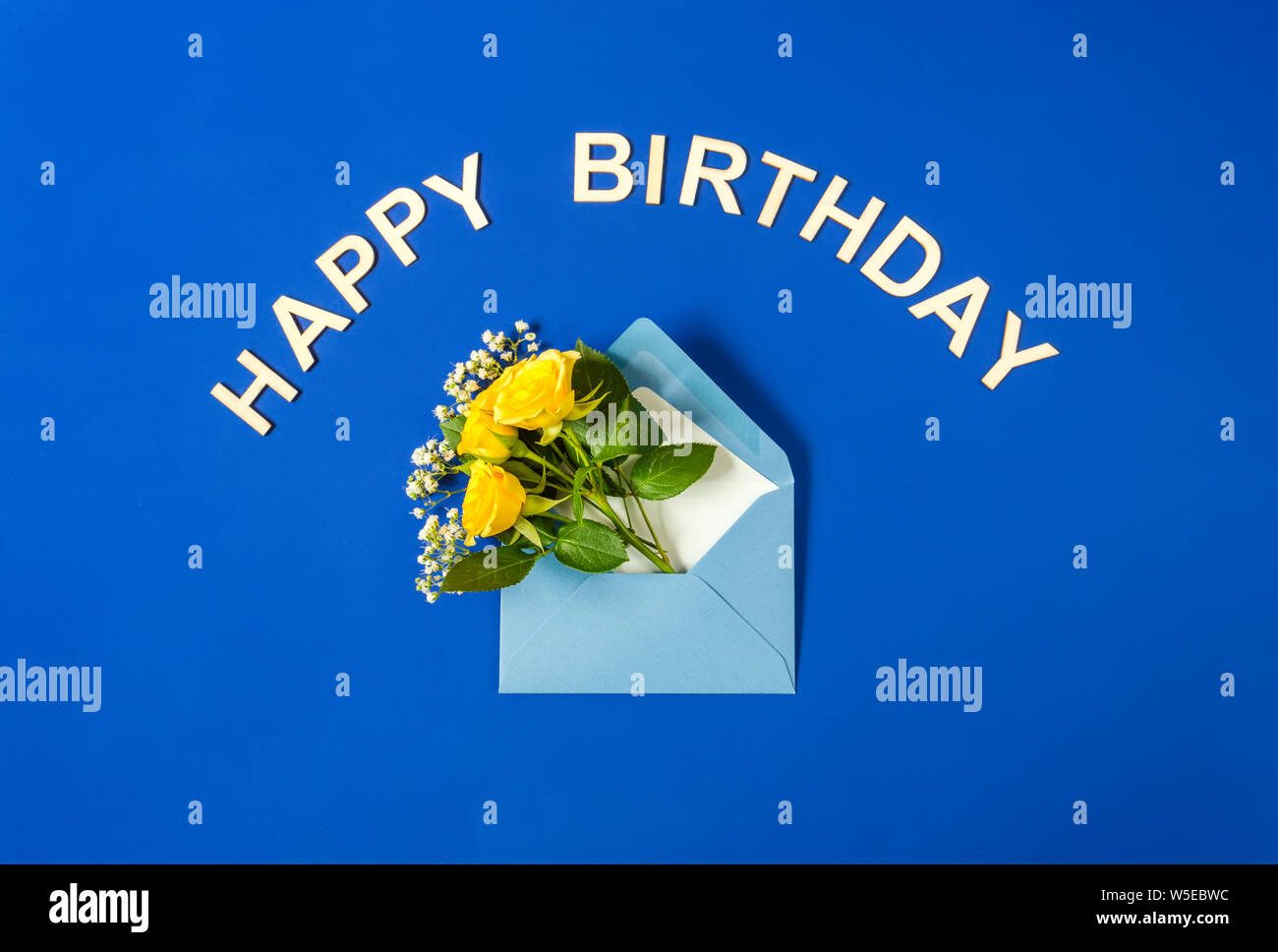 Gelbe Rosen und weißen Gypsophila in blauen Umschlag close-up auf blauem Hintergrund. Text Happy Birthday, Holz- briefe. Ansicht von oben, flach. Vorlage für Stockfoto