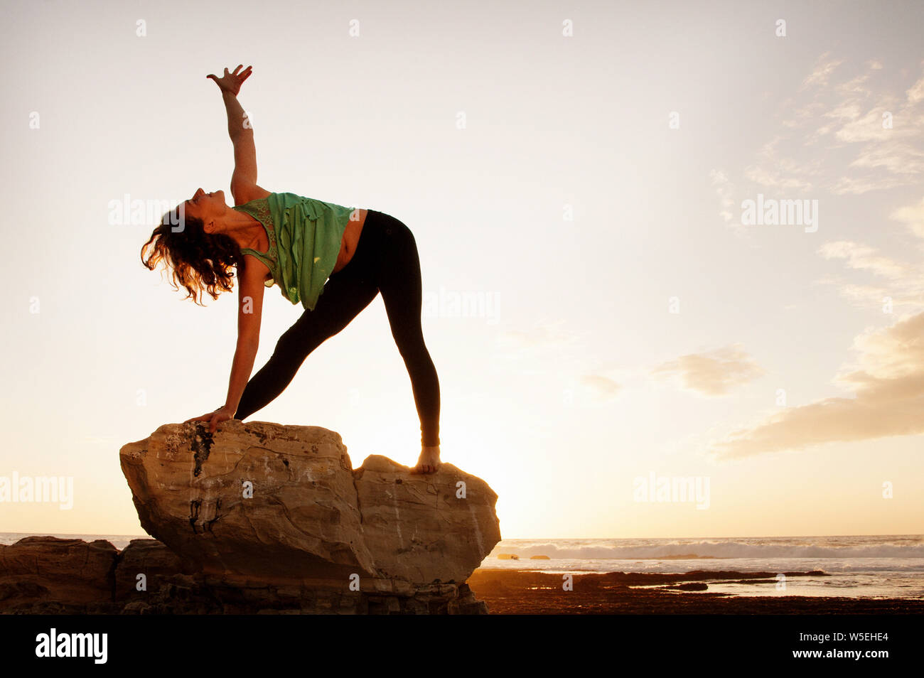 Yoga Frau in Dreieck pose am Strand bei Sonnenuntergang auf einem großen Stein. Stockfoto