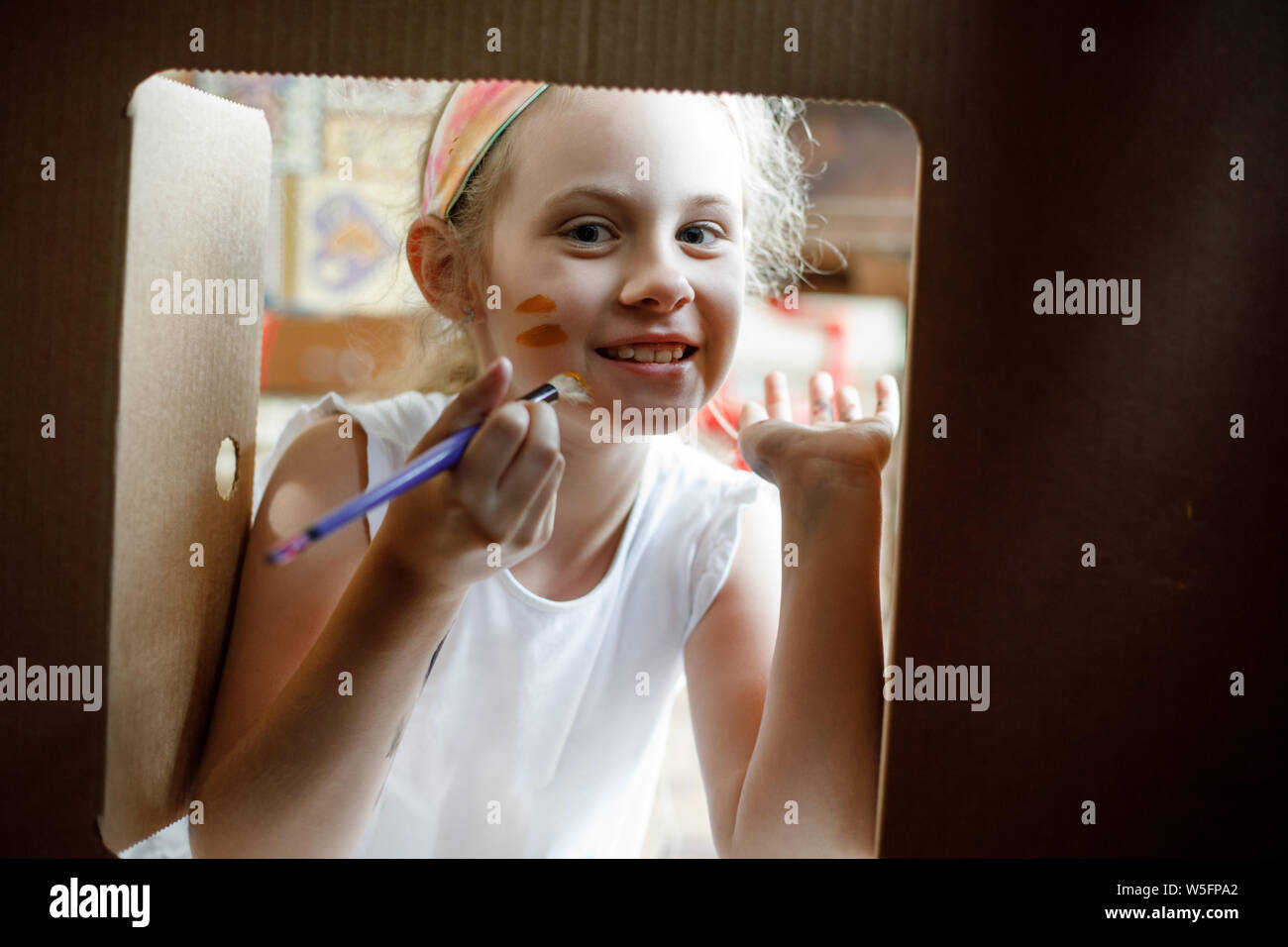 Happy girl Farben auf ihrem Gesicht schaut durch das Fenster der Pappe Haus. Stockfoto