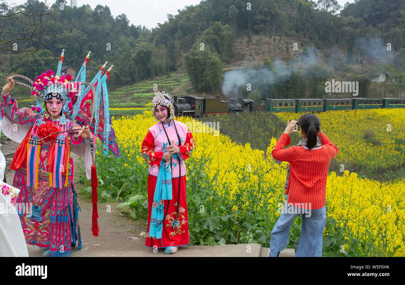 Touristen in Trachten und Sichuan Oper Kostümen stellen neben einem Zug, der durch eine Raps Feld in voller Blüte in Leshan Stadt, Stockfoto