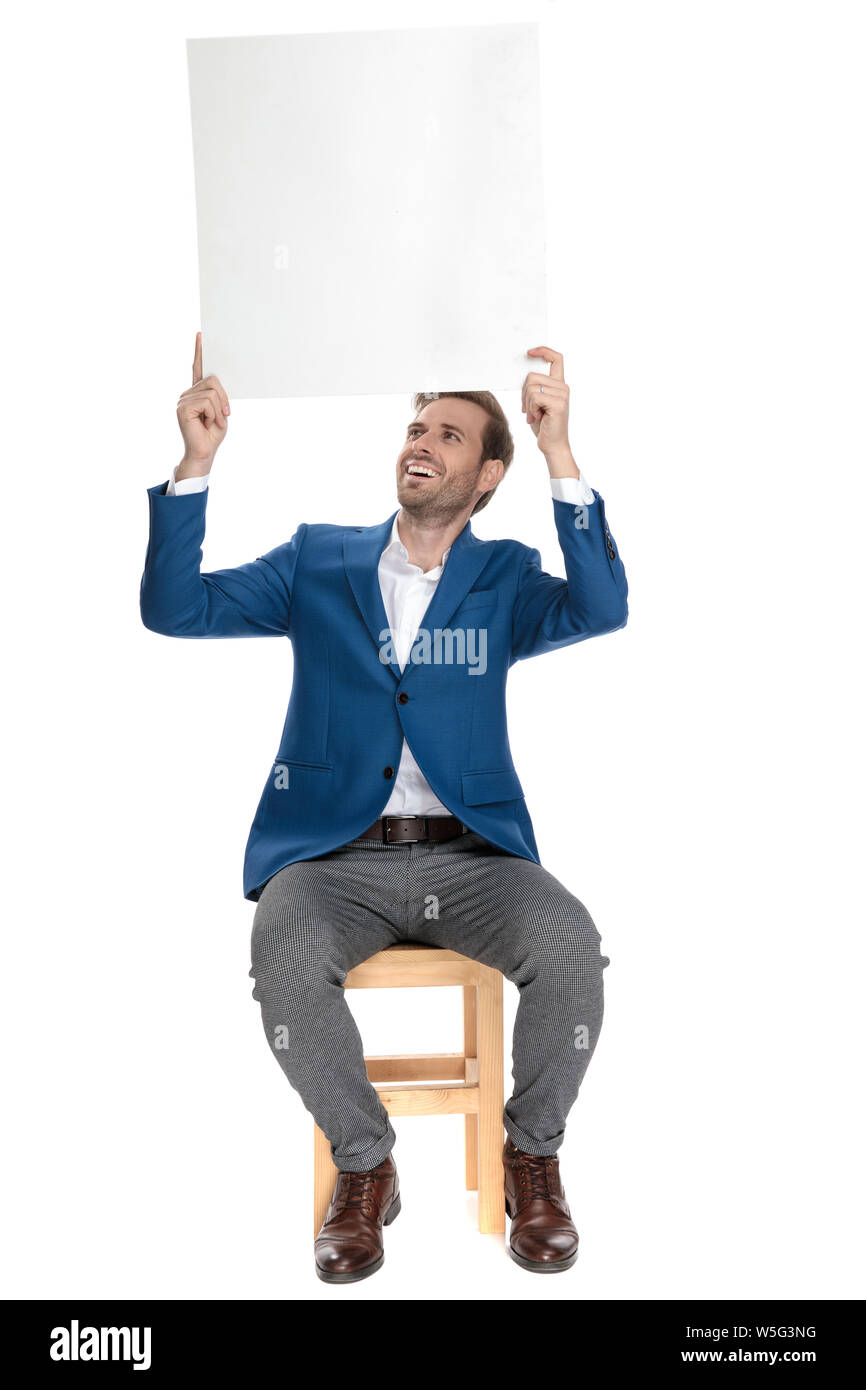 Hoffnungsvoll lässiger Kerl an der Anschlagtafel er über seinem Kopf hält beim Sitzen auf einem Stuhl und im Anzug auf weißem Studiohintergrund lachen Stockfoto