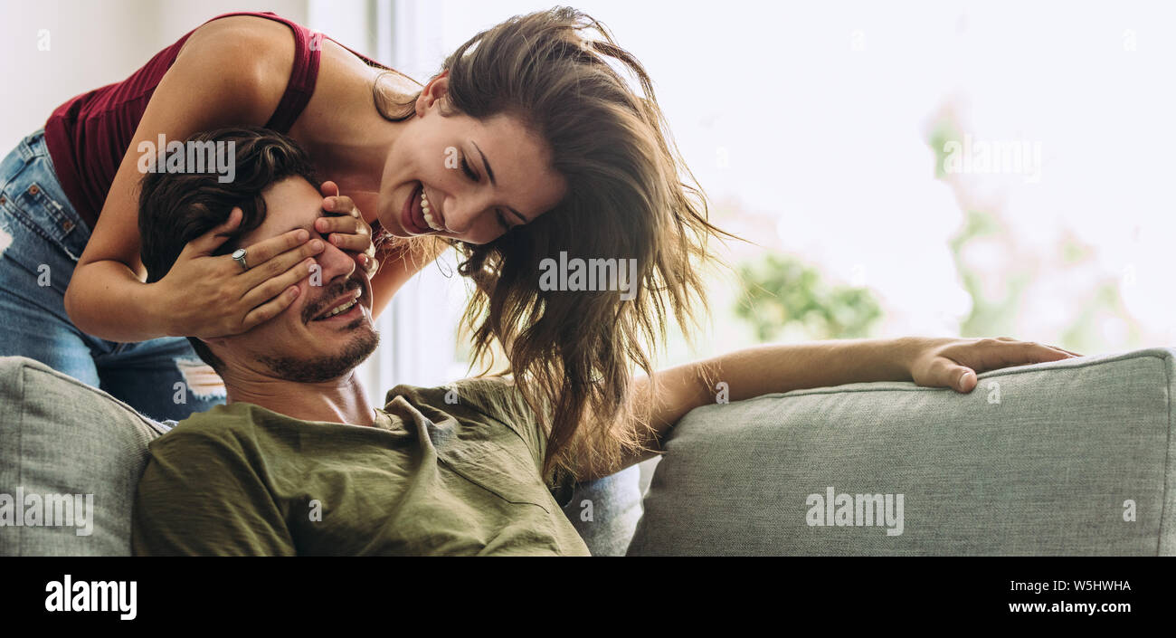 Lächelnden jungen Frau die Augen ihres Partners sitzen auf einem Sofa. Junge Frau überraschend Mann im Wohnzimmer zu Hause. Stockfoto
