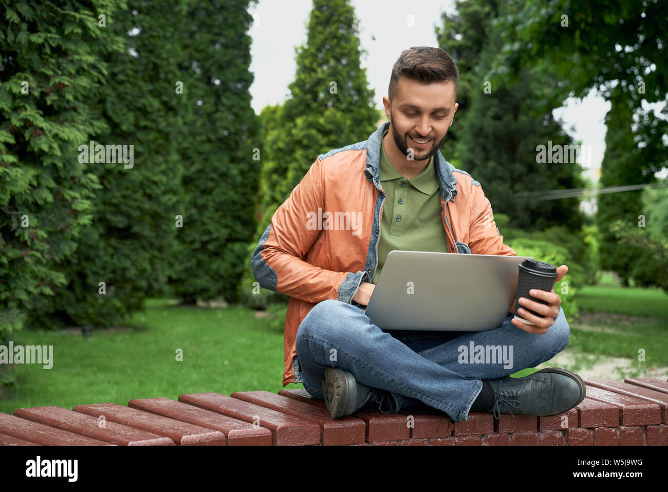 Schöne, stilvolle Mann auf Bank aus Holz im Garten, mit Laptop. Schüler Sitzen mit gekreuzten Beinen, die eine neue Tasse Kaffee, auf Laptop, lächelnd. Stockfoto