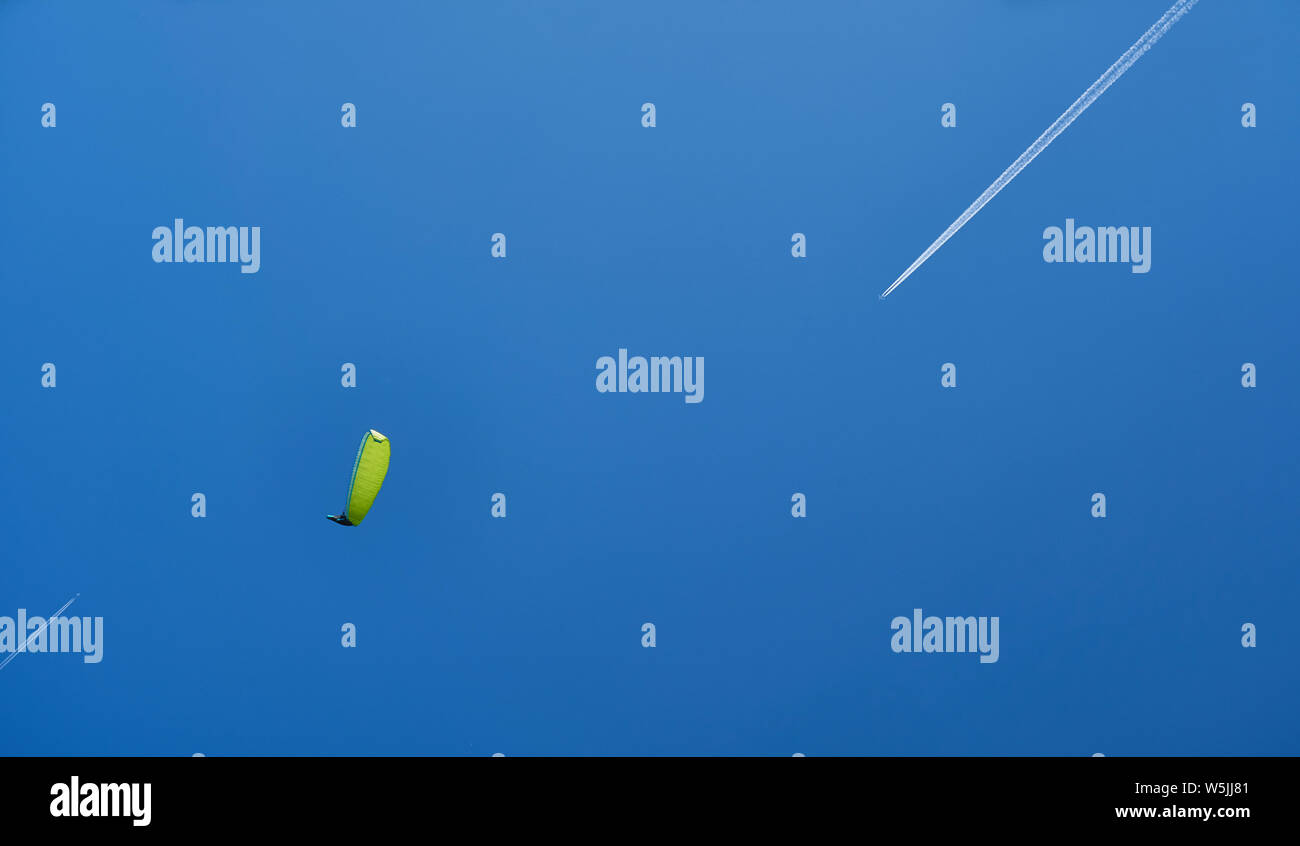 Ein Gleitschirm mit einem gelb-grün parashute und ein Flugzeug verlassen eine weiße Spur auf den blauen Himmel. Stockfoto