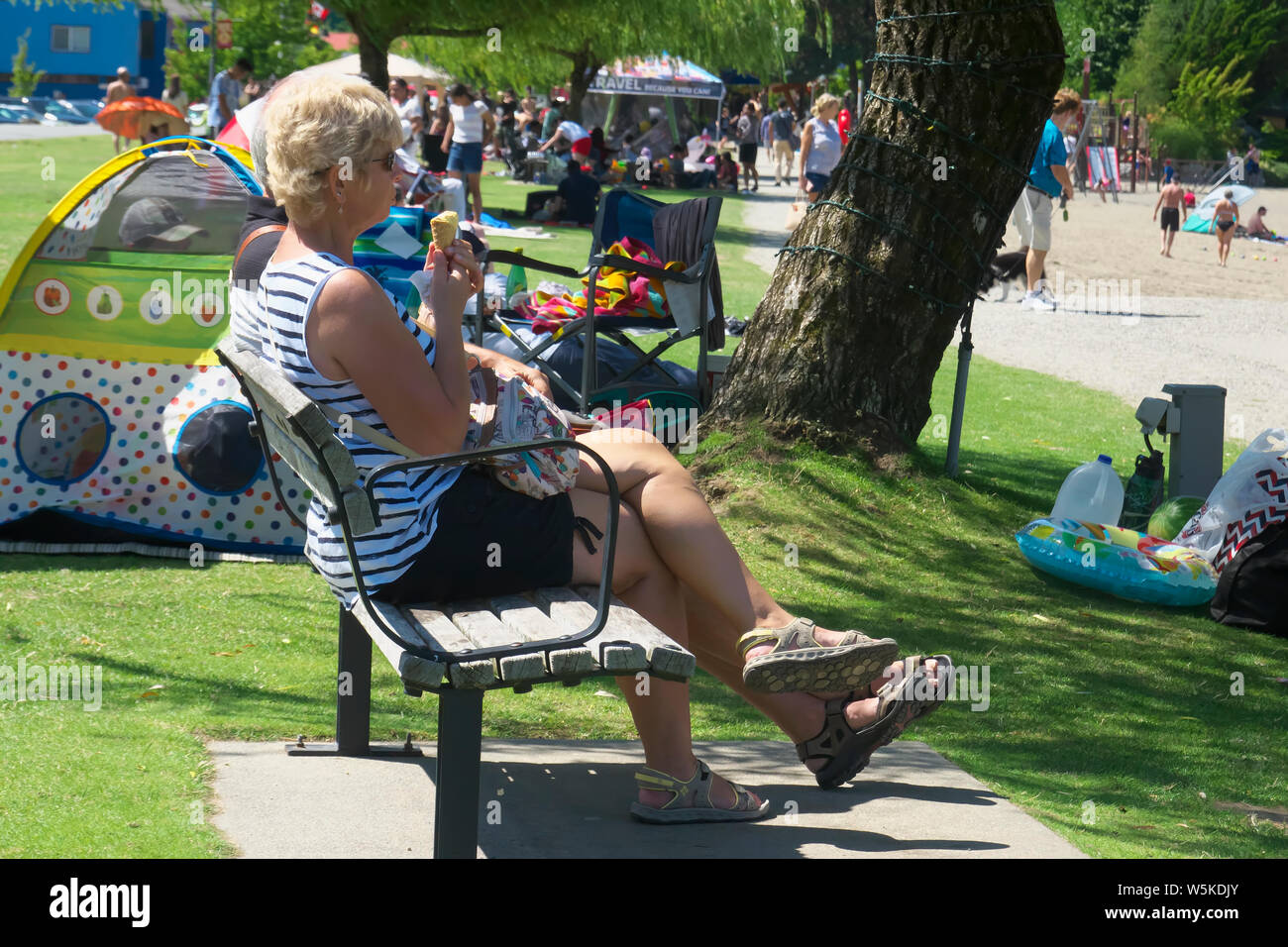 Blonde Frau auf einer Bank sitzen genießen ein Eis mit vielen Menschen im Hintergrund in Harrison Hot Springs Juli 2019. Stockfoto