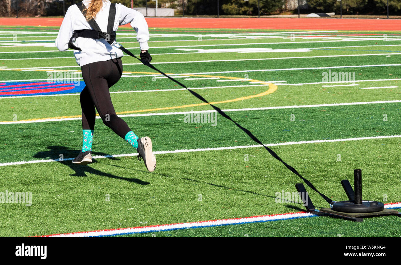 Ein High School Mädchen läuft weg von der Kamera ziehen einen Schlitten mit Gewicht über ein grünes Feld mit schwarzen spandex und grauen Sweatshirt. Stockfoto