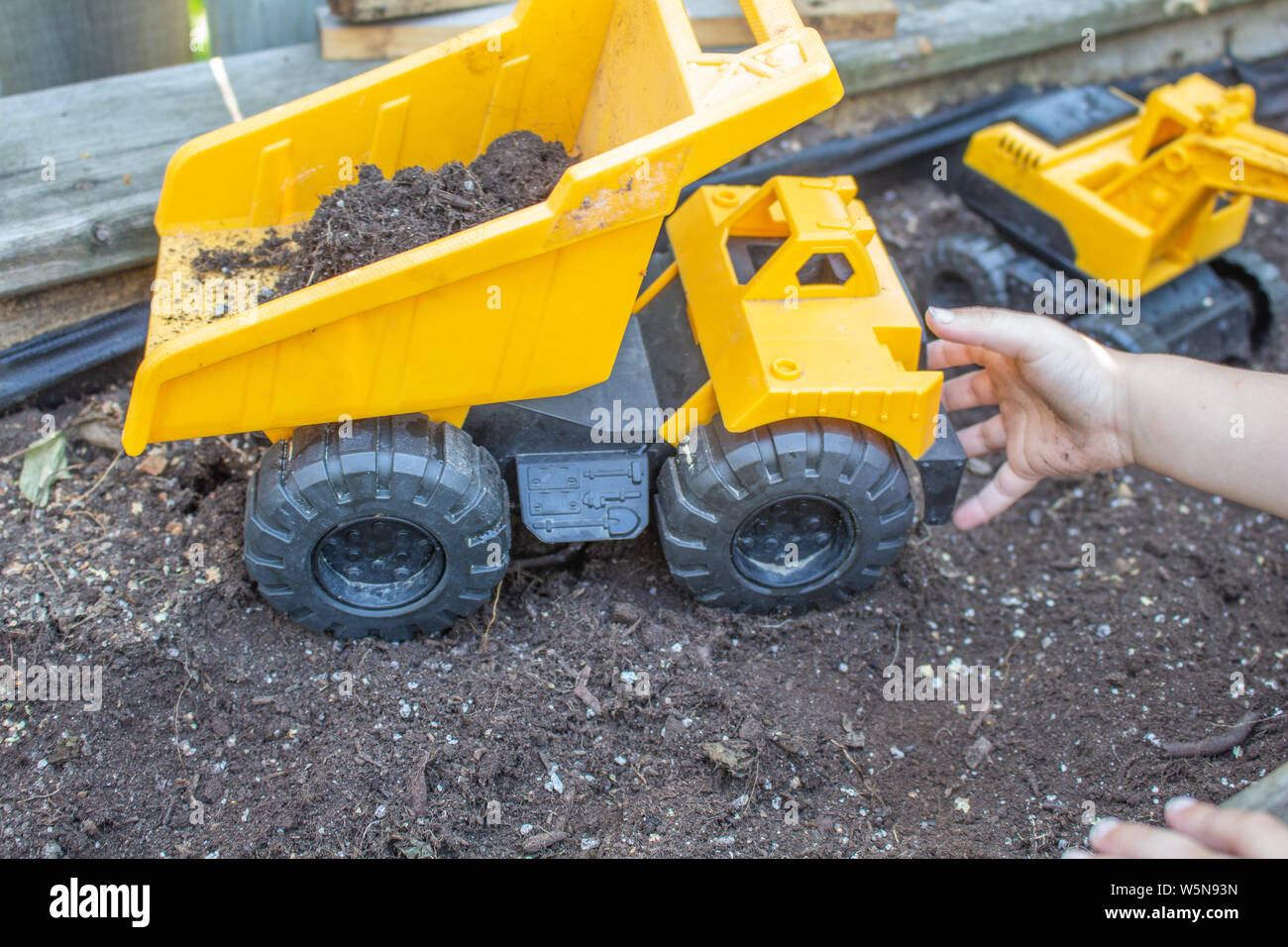 Die Hand eines jungen Kindes erreicht heraus für ein Spielzeug Kipper, sitzen auf dem Boden der einen erhöhten Blumenbeet. Stockfoto