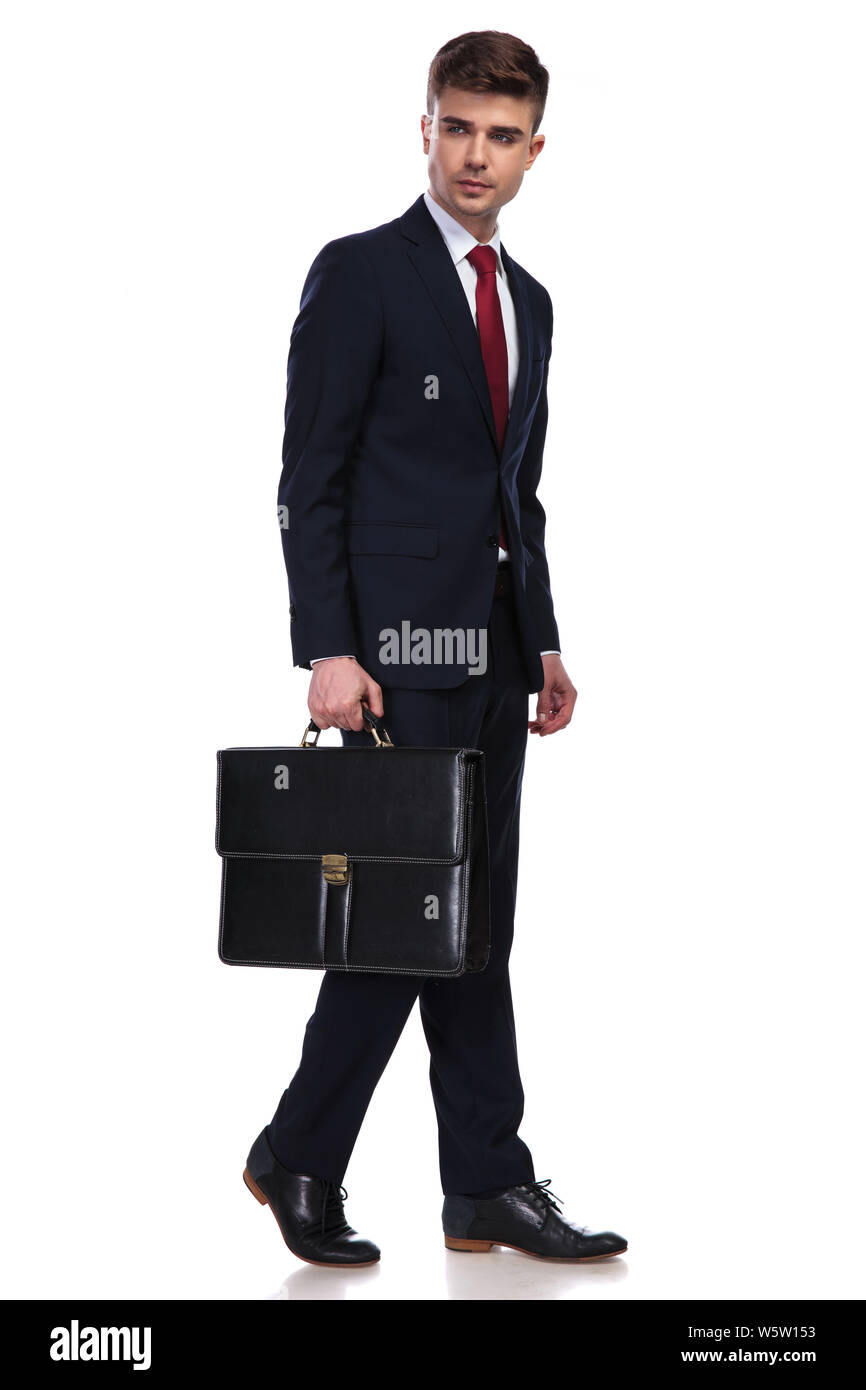 Wandern Geschäftsmann schaut zur Seite, während Sie einen Koffer. Er trägt eine Marine Anzug und eine rote Krawatte, vollen Körper Bild Stockfoto