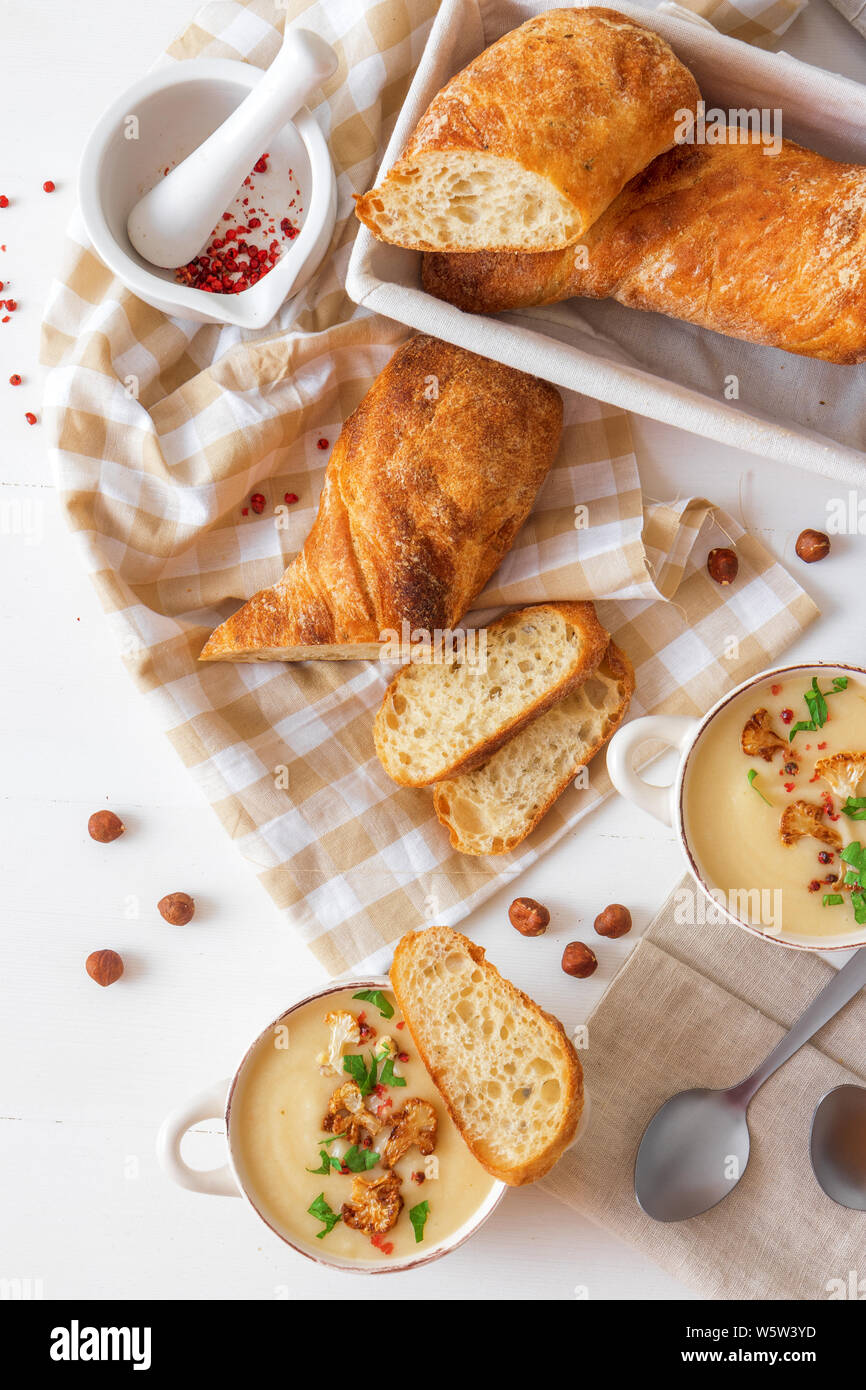 Weiße Holztisch mit 2 Schalen von Blumenkohl Suppe mit zerquetschte Haselnüsse, roter Pfeffer, glatte Petersilie Kräuter verschönert, Schweizer twisted Brot, braten Stockfoto