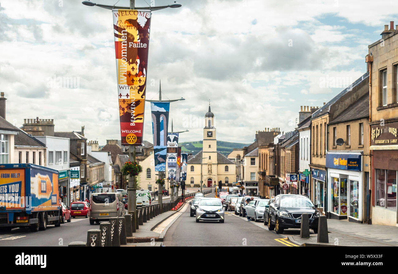 Lanark High Street mit ihren Geschäften, Fahrzeuge und Banner, die Geschichte der Stadt. In mehreren Sprachen Willkommen. Lanarkshire, Schottland Stockfoto