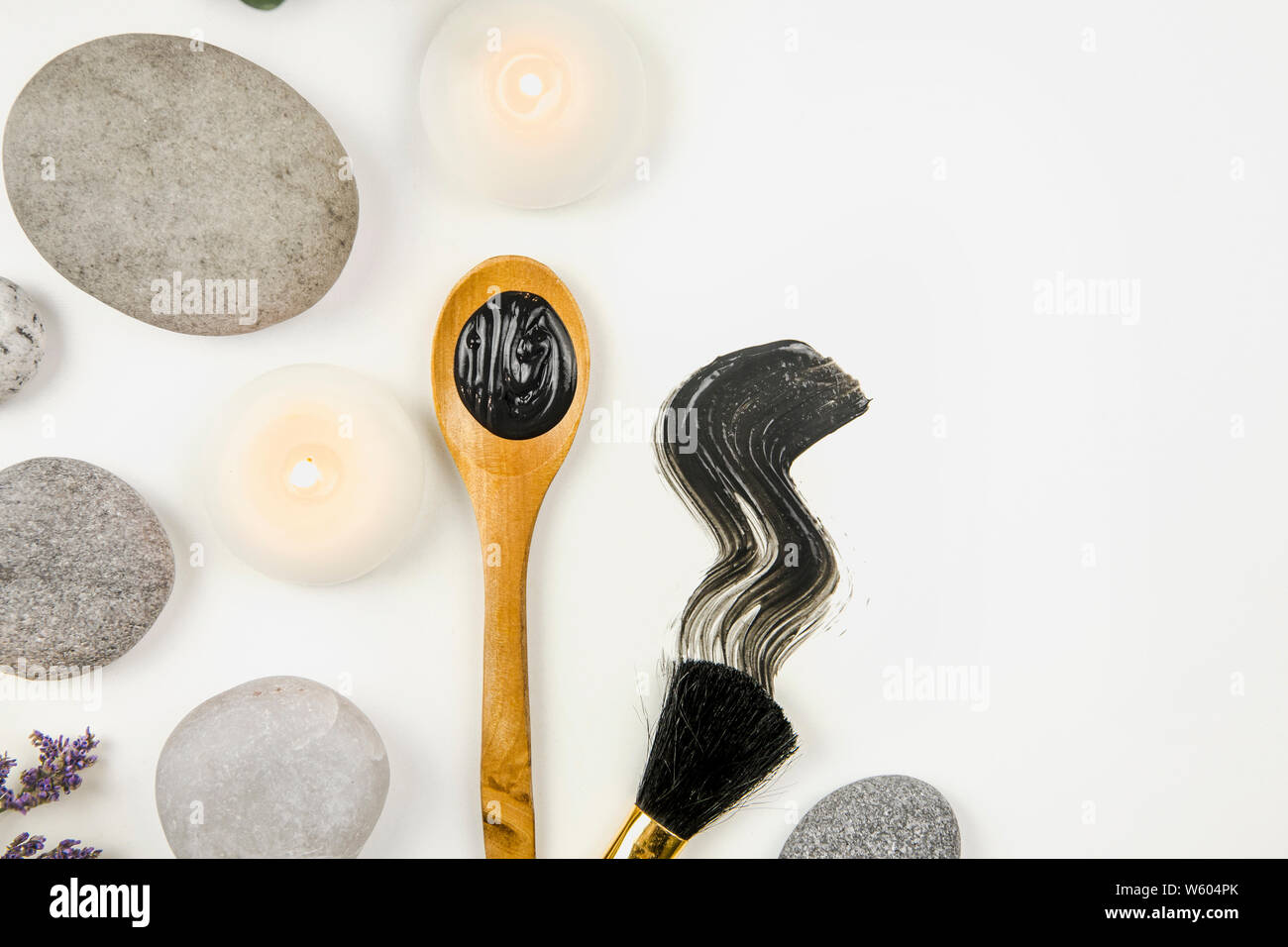 Flach auf Schlamm Holzkohle Maske auf Holzlöffel und Abstrich auf weißem Hintergrund, mit Schönheit Pinsel und flache Meer Steine und spa Kerzen umgeben. Ro Stockfoto