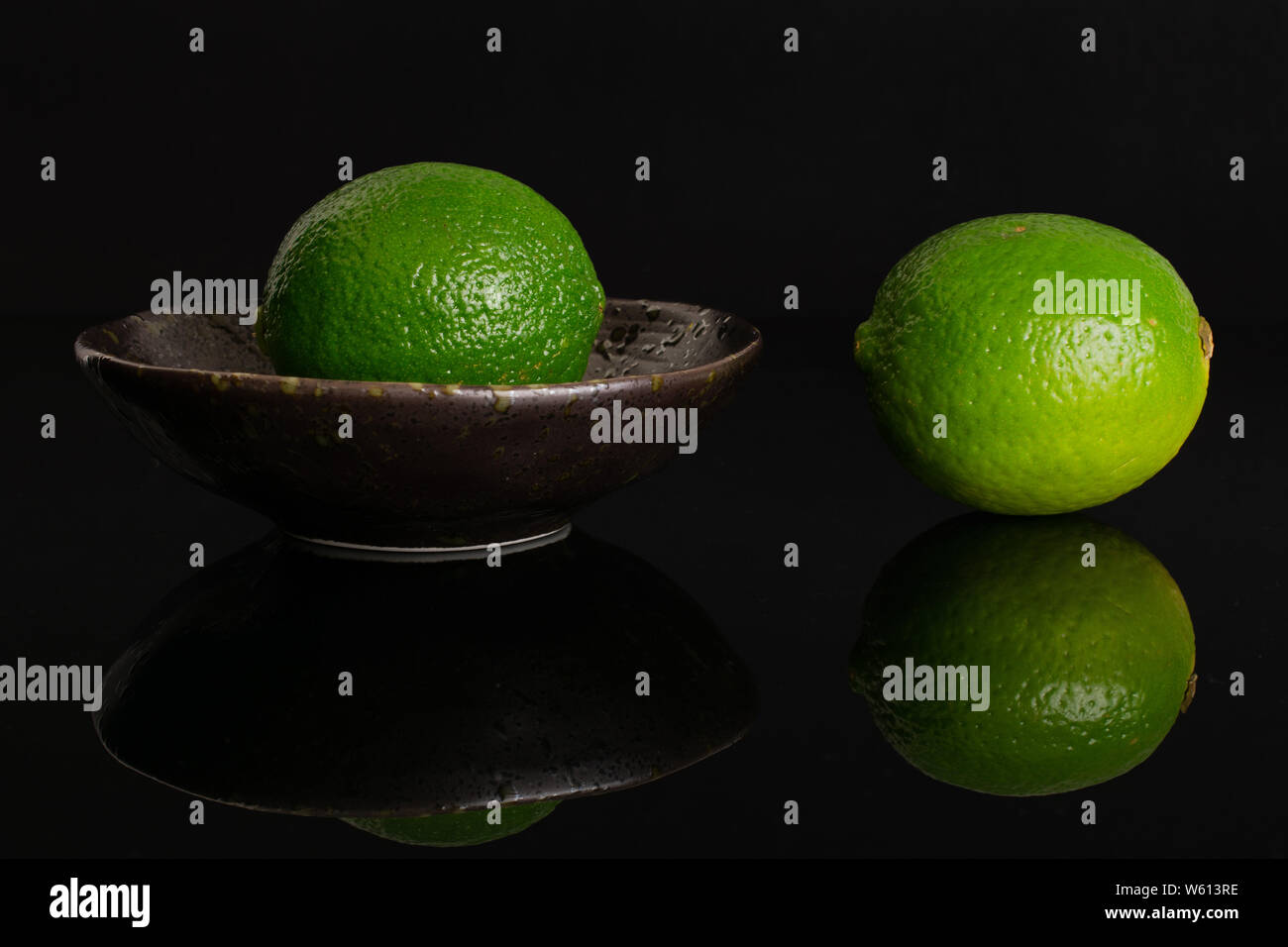 Gruppe von zwei ganze Frische grüne Kalk ist man beiseite und der andere ist in einem dunklen Keramik Schüssel auf schwarzem Glas isoliert Stockfoto