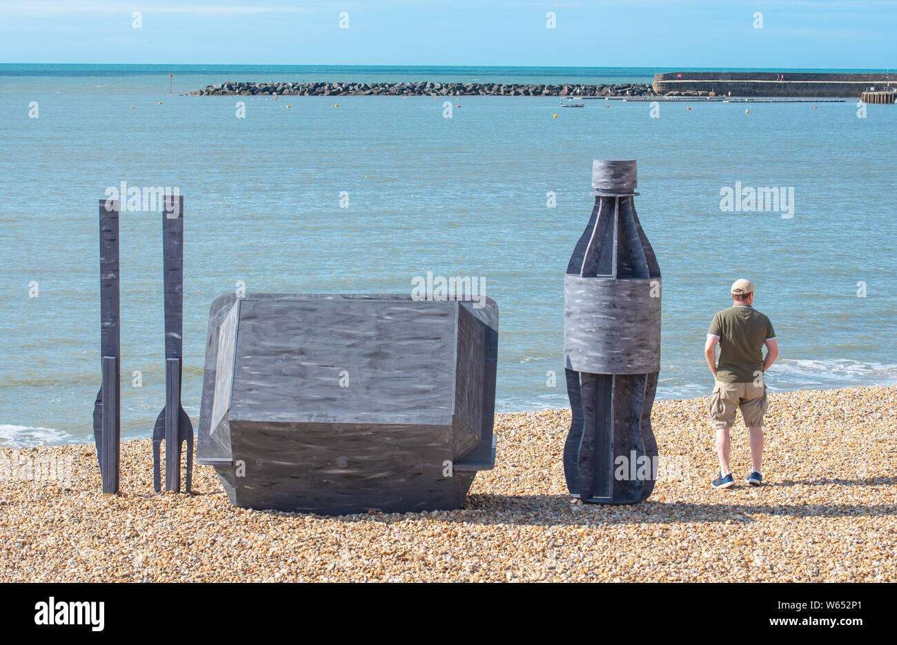 Lyme Regis, Dorset, Großbritannien. 31. Juli 2019. UK Wetter: ein Mann bewundert die Aussicht aus einem riesigen Wurf Skulptur von Takeaway Verpackung, die auf dem malerischen Strand an der Küste von Lyme Regis errichtet wurde. Bestehend aus 8 Fuß hoch Kunststoff Trinkflasche, Besteck 6 Fuß hoch und einem Chip/burger Box 4 x 4 Fuß die Skulpturen die Bemühungen der Wurf frei Küste und Meer und Partner bei der Förderung ökologisch verantwortungsvolle Entscheidungen zu fördern. Es ist zu hoffen, dass die vorübergehende Installation von architektonischen Designer Jack Hebeny Leute anregen ihren Müll mit nach Hause zu nehmen. Gutschrift: Stockfoto