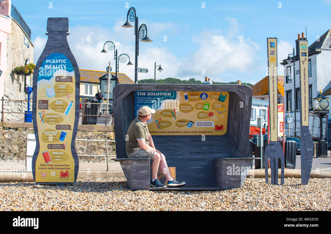 Lyme Regis, Dorset, Großbritannien. 31. Juli 2019. UK Wetter: ein Mann bewundert die Aussicht aus einem riesigen Wurf Skulptur von Takeaway Verpackung hat hat am malerischen Strand errichtet worden in den Badeort Lyme Regis. Bestehend aus 8 Fuß hoch Kunststoff Trinkflasche, Besteck 6 Fuß hoch und einem Chip/burger Box 4 x 4 Fuß die Skulpturen die Bemühungen der Wurf frei Küste und Meer und Partner bei der Förderung ökologisch verantwortungsvolle Entscheidungen zu fördern. Es ist zu hoffen, dass die vorübergehende Installation von architektonischen Designer Jack Hebeny ermutigen wird, die Besucher ihren Müll mit nach Hause zu nehmen. Gutschrift: Stockfoto