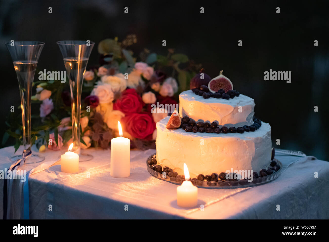 Hochzeit Dekorationen. Kuchen in weiße Glasur mit einer Einrichtung der Blaubeeren und Feigen auf dem Tisch am Abend mit Gläsern, Kerzen und eine Hochzeit bouq Stockfoto