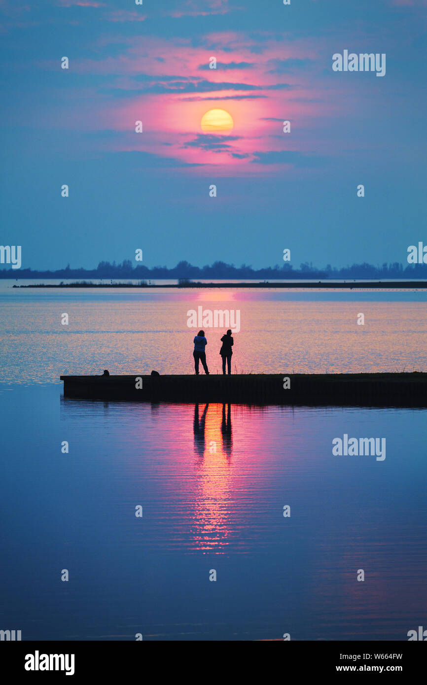 Ein romantischer Sonnenuntergang mit den Menschen auf einem Steg am See gemeinsam beobachten, wie die Sonne auf einen bunten und warmen Sommern Abend-lifestyle-Bild Stockfoto