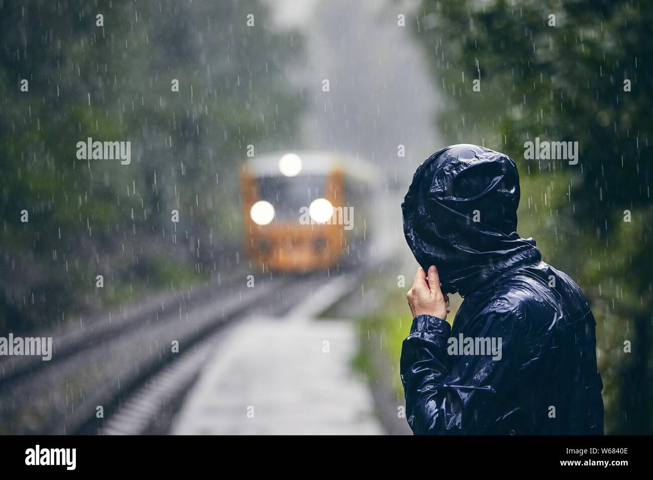 Mann in Durchnäßte Jacke steht auf Plattform der Bahnhof gegen Zug bei schwerem Regen. Stockfoto