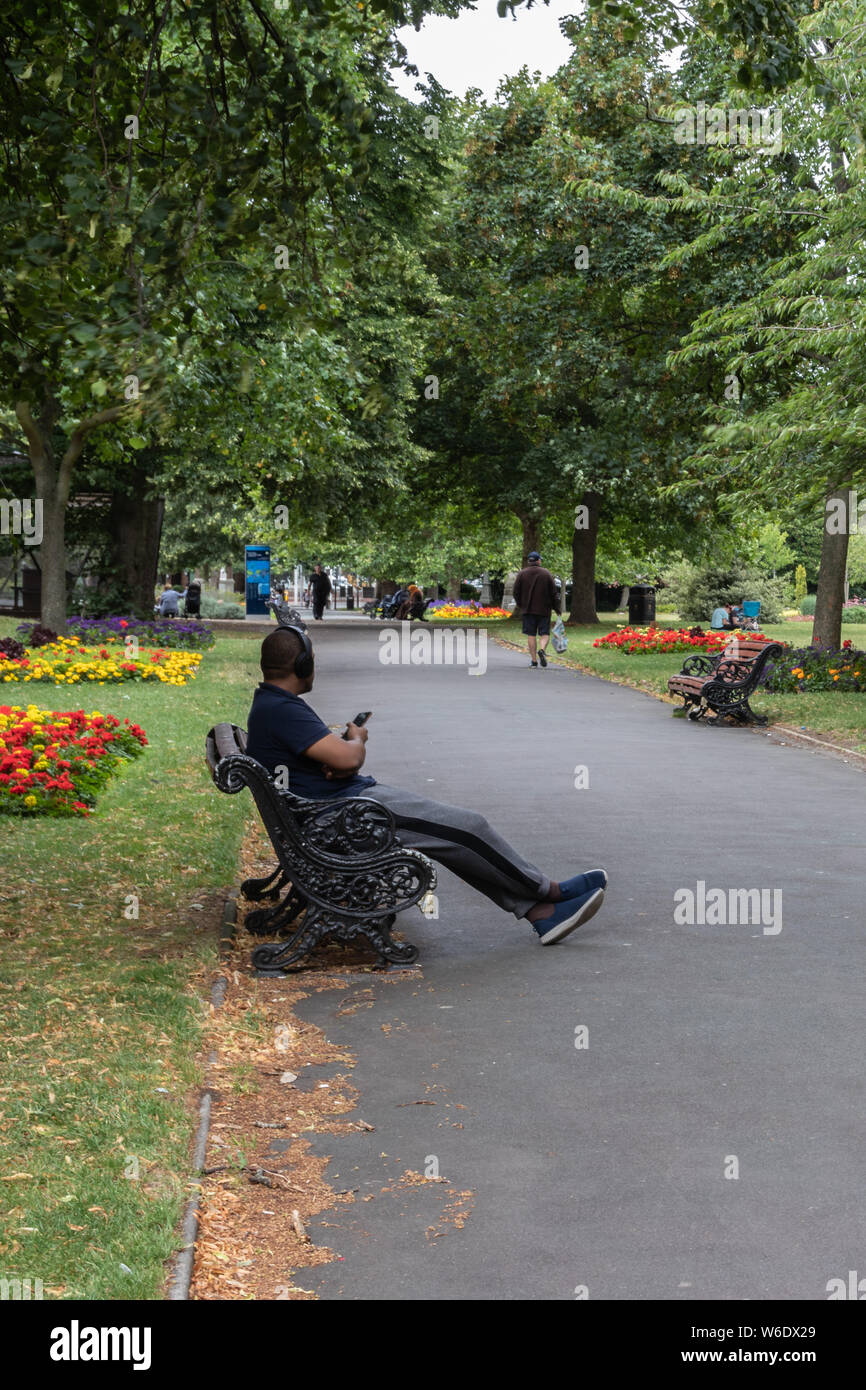 Ein schwarzer Mann saß auf einer Bank im Park mit Kopfhörern sein Handy oder Telefon verkaufen Stockfoto
