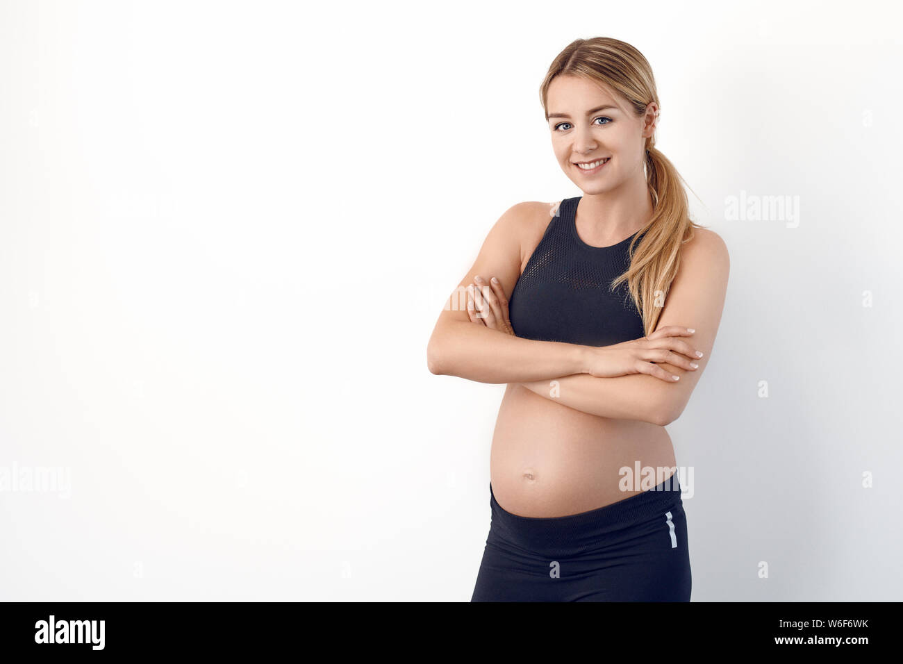 Glücklichen gesunden jungen schwangeren Frau in Schwarz Sportbekleidung auf den Hüften in die Kamera schaut mit einem strahlenden Lächeln Stockfoto