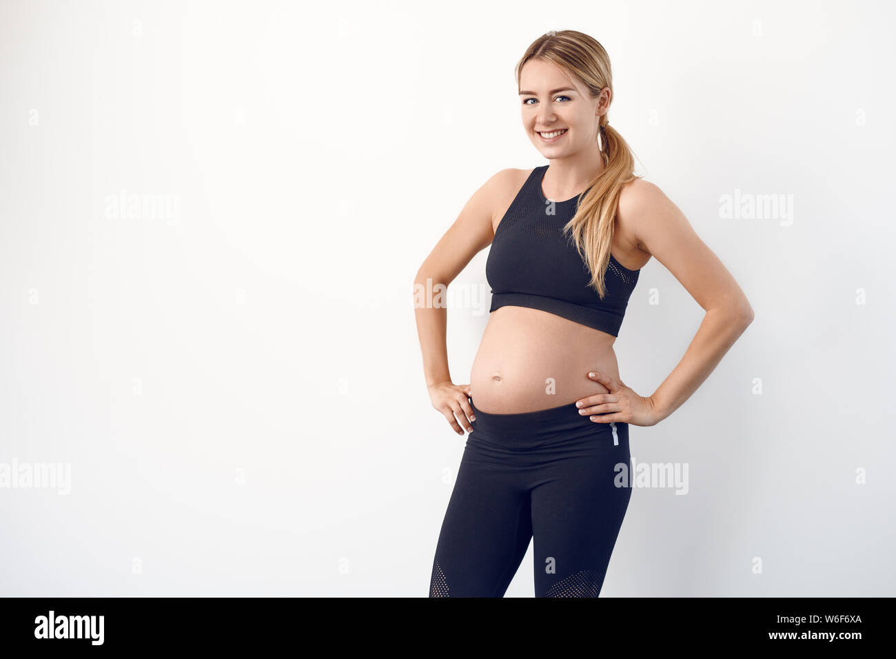 Glücklichen gesunden jungen schwangeren Frau in Schwarz Sportbekleidung auf den Hüften in die Kamera schaut mit einem strahlenden Lächeln Stockfoto