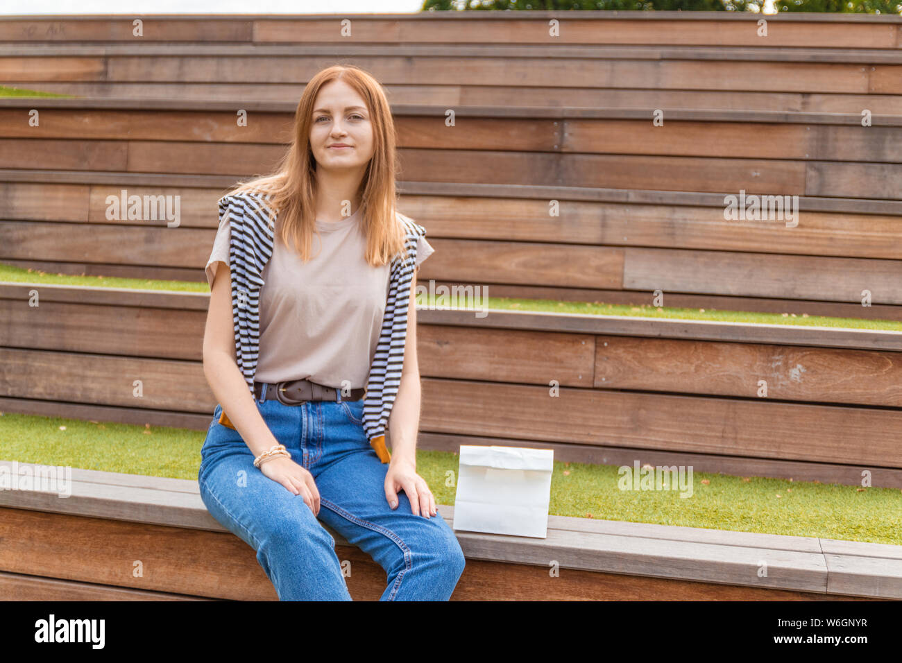 Lächelnde blonde Schüler Mädchen in T-Shirt und Blue Jeans sitzt auf einer Holzbank mit einer Doggy Bag auf einen sonnigen Tag im Park. Mittagspause. Leer Stockfoto