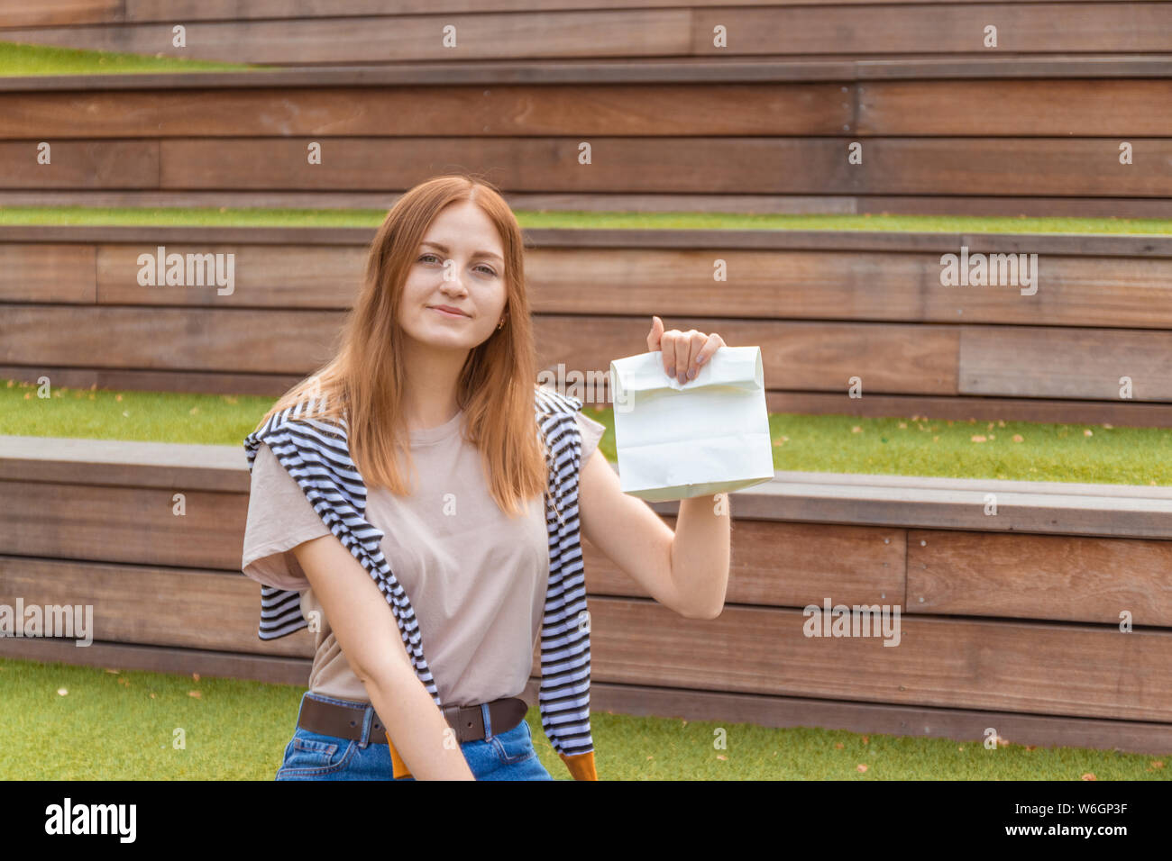 Lächelnde blonde Schüler Mädchen in T-Shirt und Blue Jeans sitzt auf einer Holzbank mit einer Doggy Bag auf einen sonnigen Tag im Park. Mittagspause. Leer Stockfoto