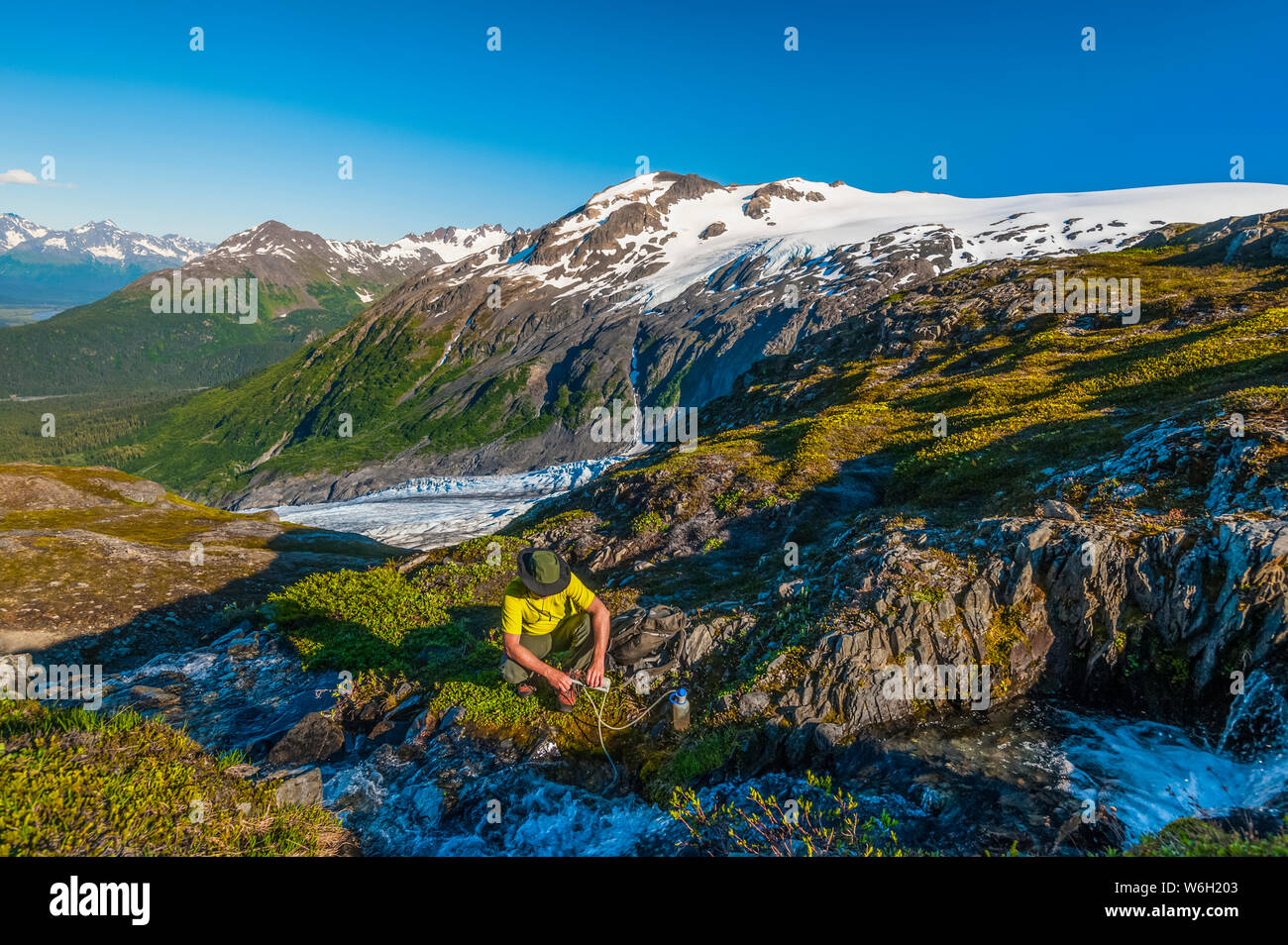 Ein Mann, der mit dem Rucksack unterwegs ist und seine Wasserflasche aus einem kleinen Bach im Kenai Fjords National Park mit dem Exit Glacier im Hintergrund auf einem sonnigen... Stockfoto