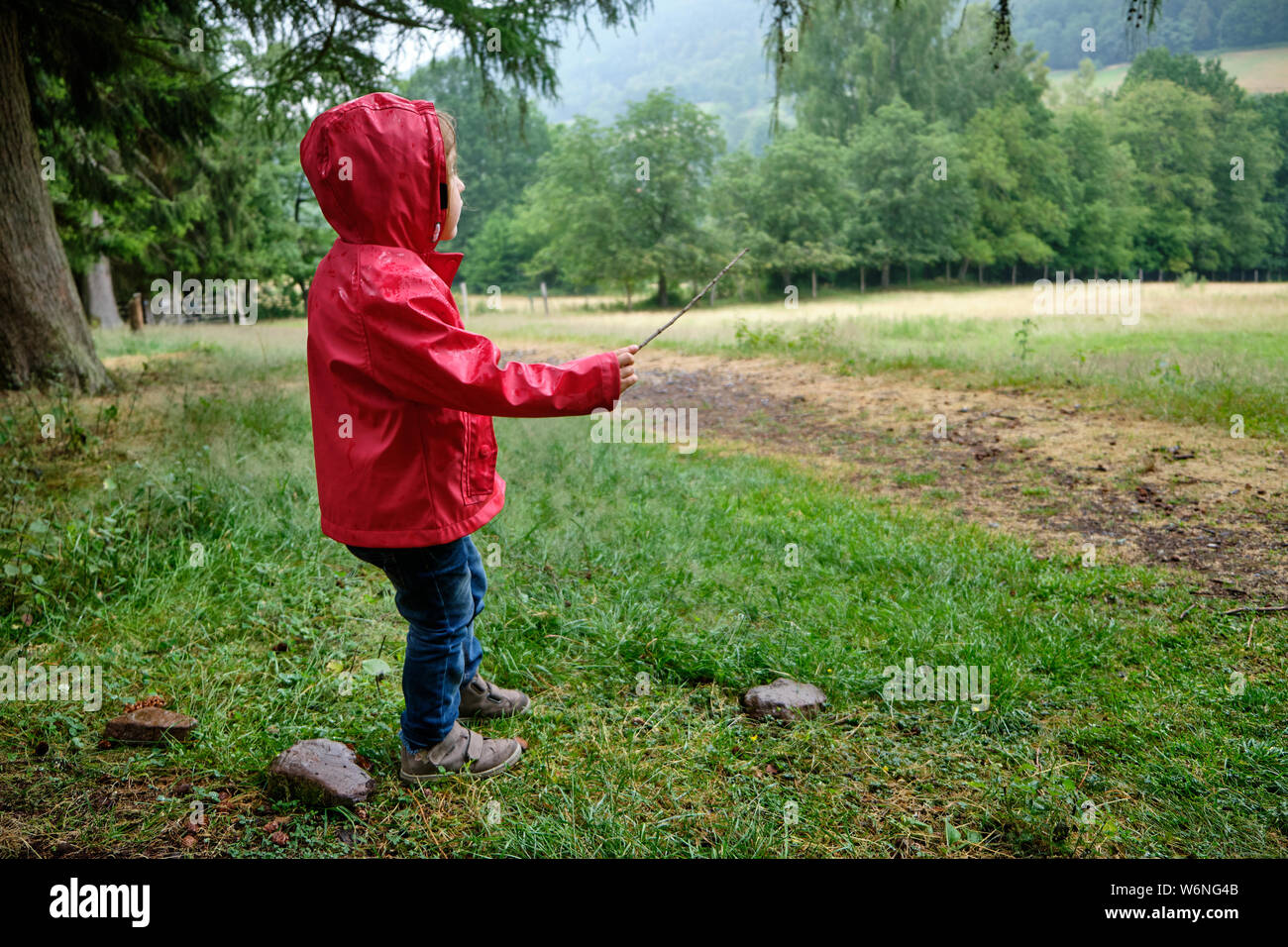 In voller Länge auf ein 3-4 Jahre altes Mädchen spielen im Freien in einer regnerischen ländliche Landschaft in Deutschland trägt eine rote Regenjacke Stockfoto