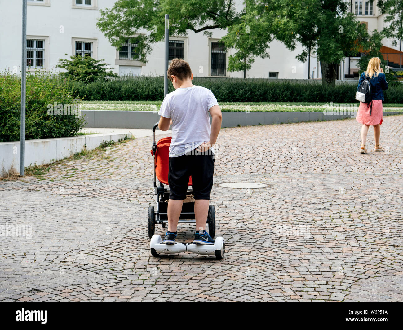 Baden-Baden, Deutschland - Apr 7, 2019: Rückansicht der faulen jungen Mann drücken Warenkorb Kinderwagen mit Baby beim Reiten eines elektrischen Hoverboard auf der gepflasterten Straßen von Baden-Baden Stockfoto