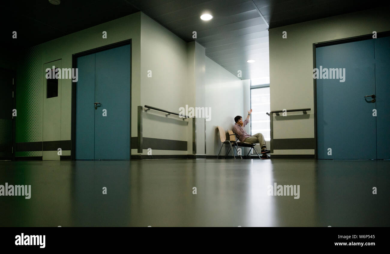 Low Angle View von einsamen Patienten sitzen auf Stuhl gestikulierte während des Sprechens auf dem Mobiltelefon im modernen Krankenhaus warten Lobby Zimmer warten auf gute oder schlechte Nachrichten von seinem Arzt Stockfoto