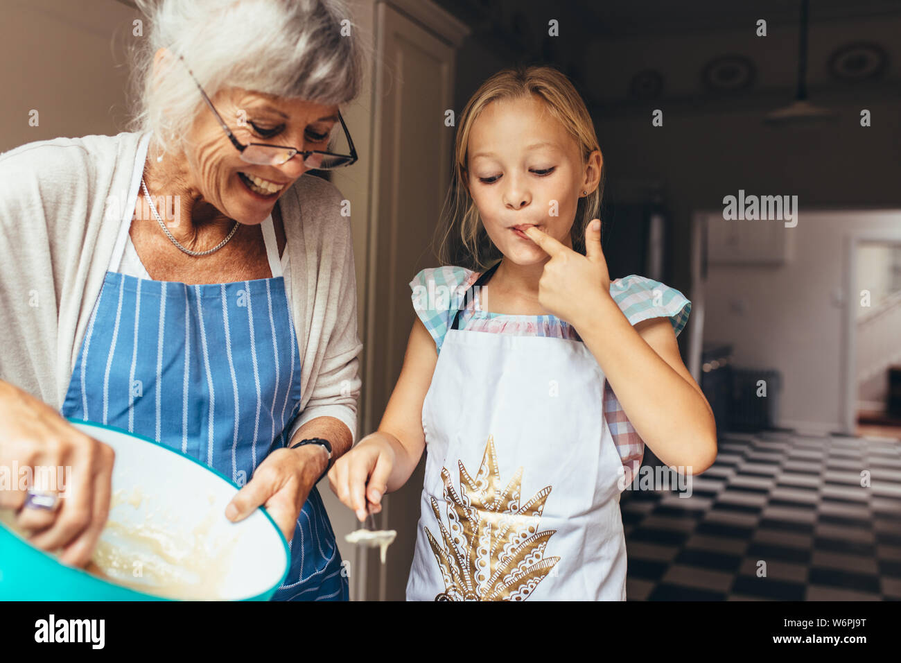 Ältere Frau in Schürze und Teig für Kuchen. Kleines Mädchen Verkostung Kuchenteig in Küche mit Großmutter. Stockfoto