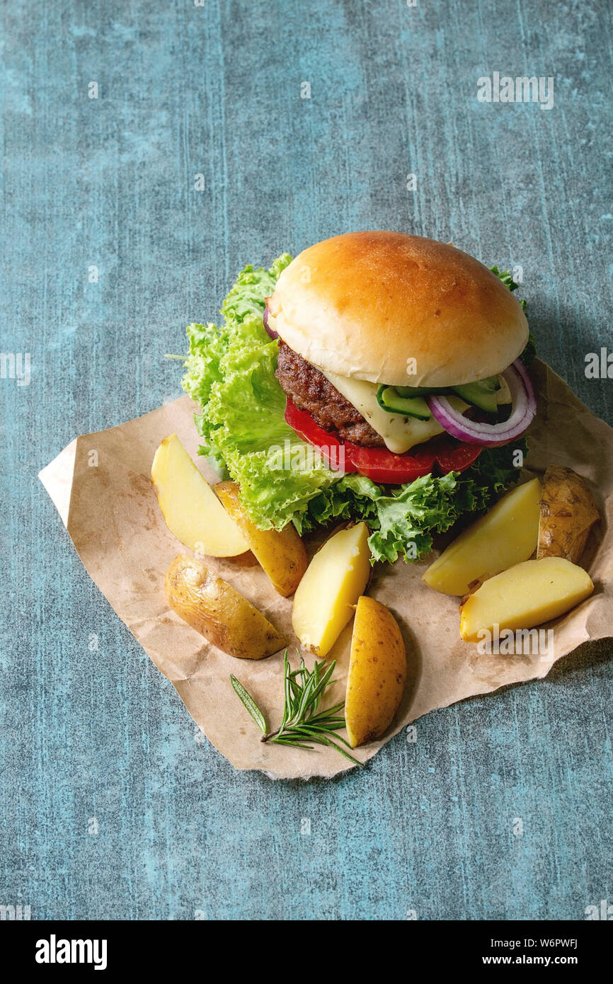 Hausgemachte Fast Food Burger classic Hamburger oder Cheeseburger mit Rindfleisch, Salat, Käse und Tomaten serviert auf Papier mit gebackenen Land Kartoffeln auf Blau te Stockfoto