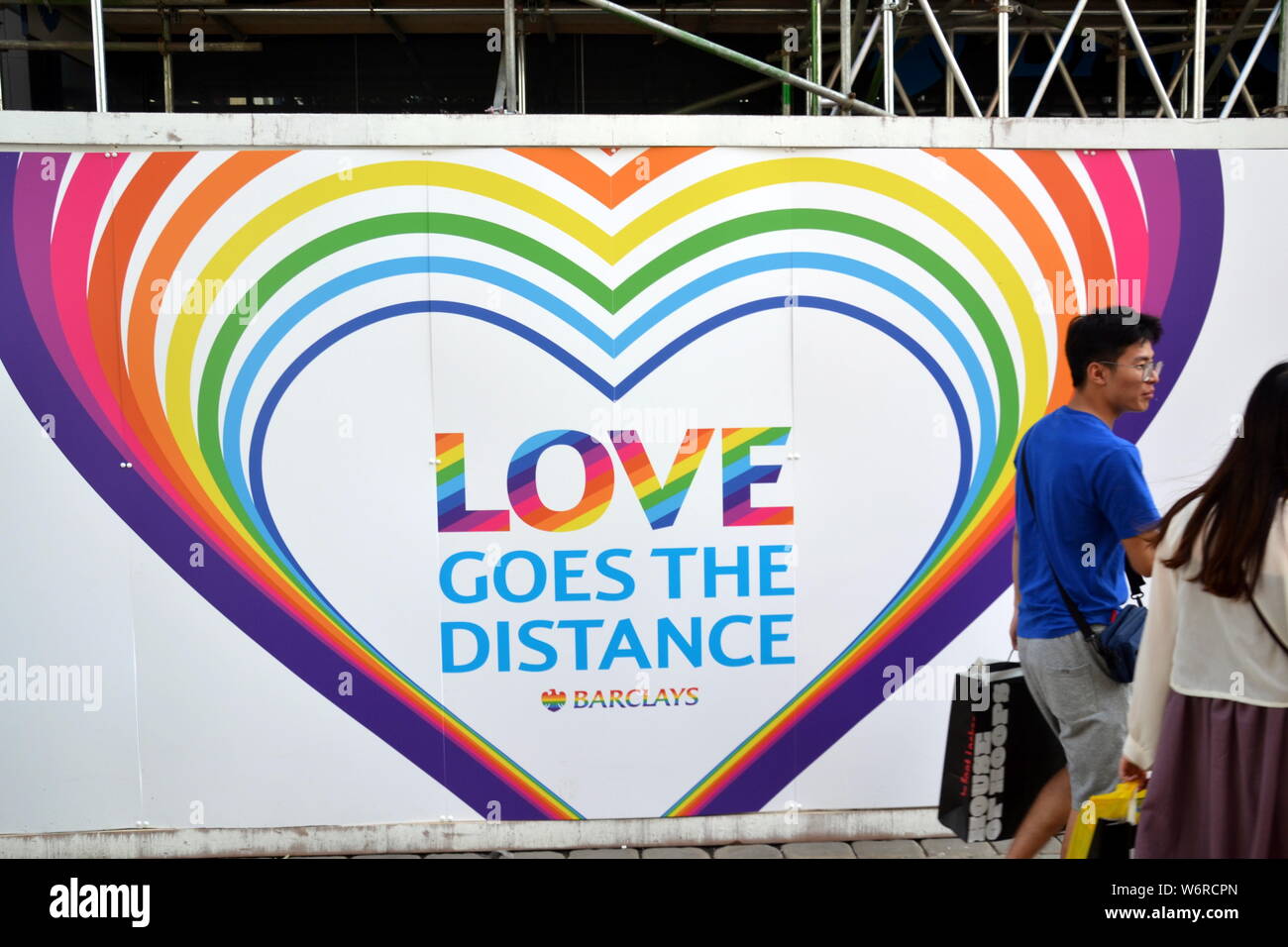 Passanten kommen während der lgbt Pride-Zeit an einem Regenbogenschild „Love Goes the Distance“ auf der Barclays Bank, Market Street, Manchester, Großbritannien, vorbei Stockfoto