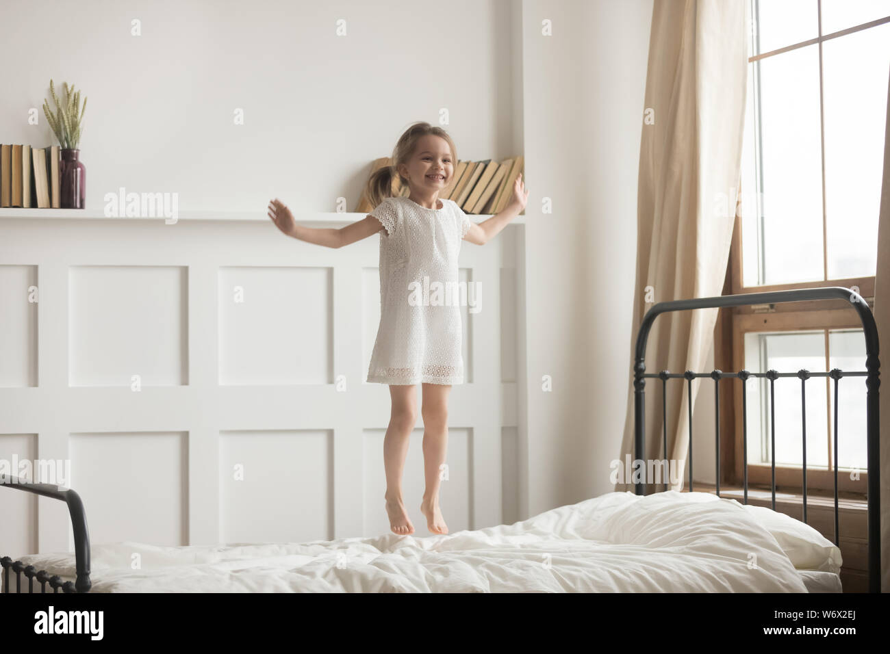 Sorglos aktiver kleiner Mädchen mit weißem Kleid springen auf Bett Stockfoto