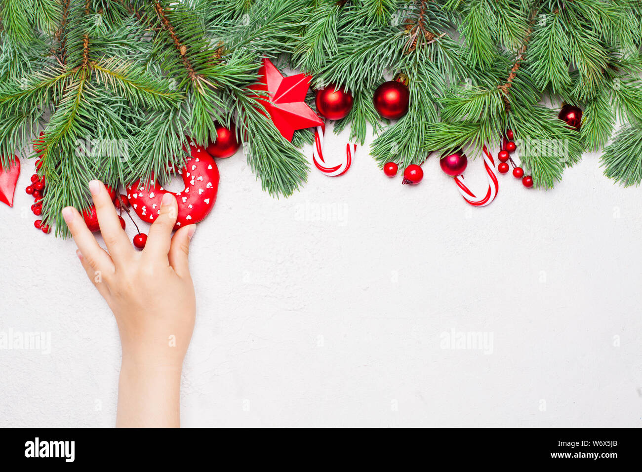 Weihnachten Konzept. Kleines Mädchen Hand mit Krapfen, Grüne Weihnachten tanne Zweig, rot Stechpalme Beeren und Kugeln auf weißen Hintergrund. Bunte Weihnachten comp Stockfoto