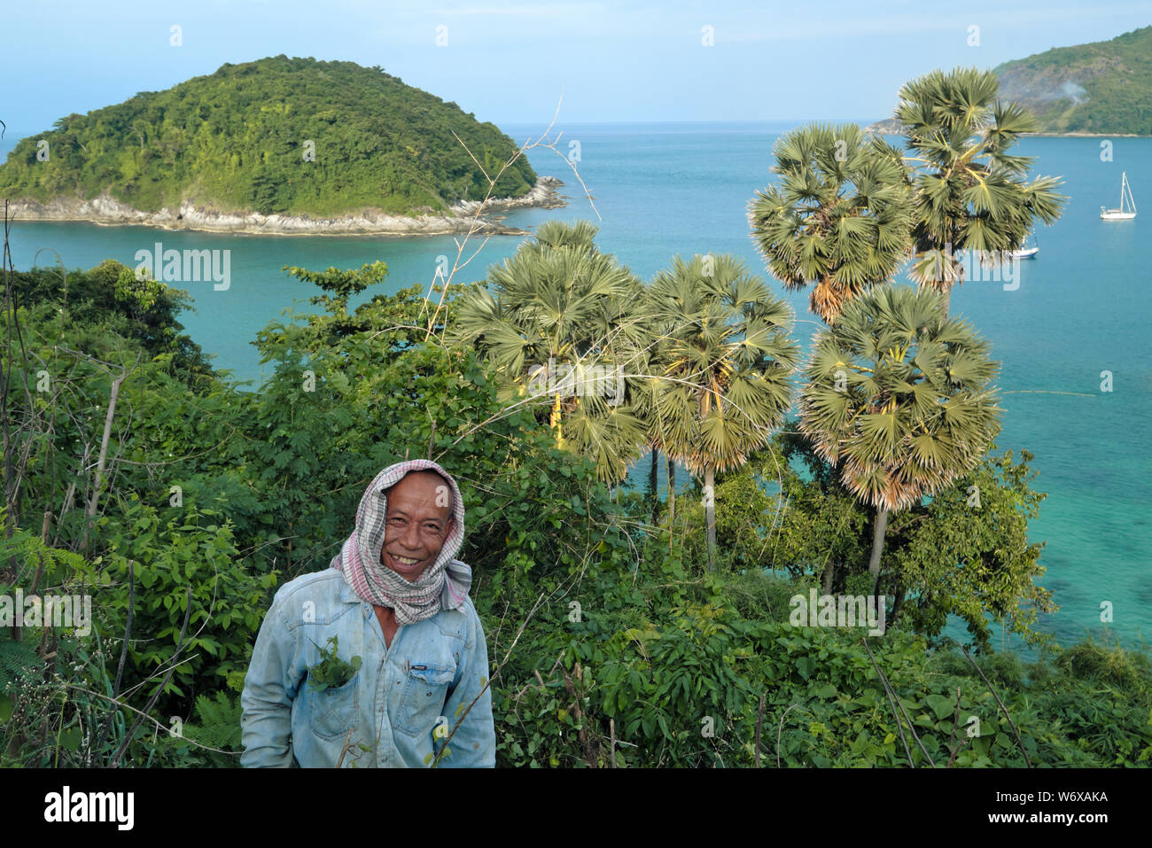 Ein Mann der Nahrungssuche für wildes Gemüse entsteht aus einem überwucherten Felsen in Ya Nui, Nai Harn, Phuket, Thailand, einer kleinen Insel und Yachten im Hintergrund Stockfoto