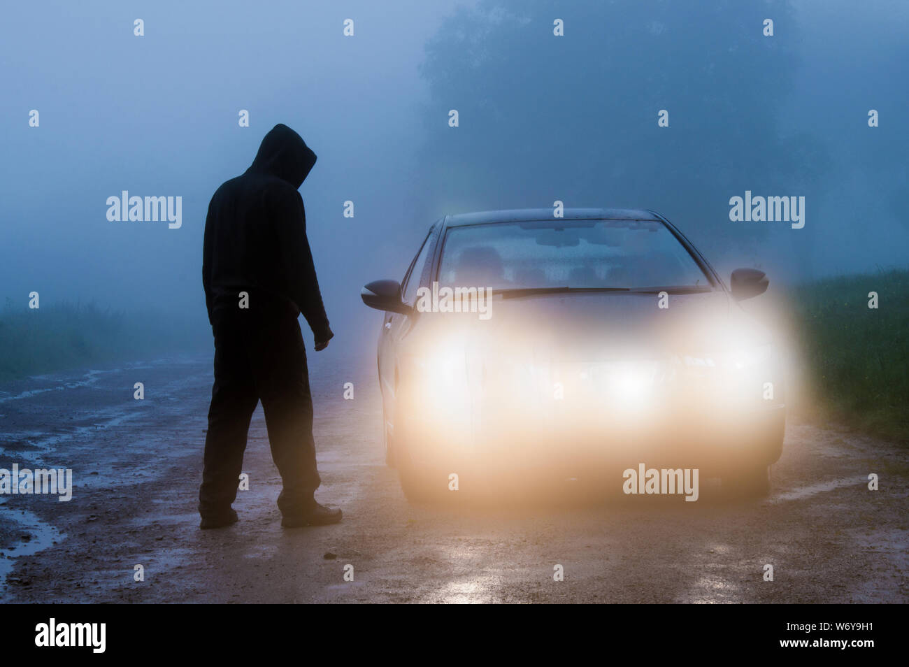 Eine bedrohliche Lone hooded Abbildung neben einem Auto auf einem nebligen spooky Straße bei Nacht. Mit dem Auto Scheinwerfer hervorgehoben. Stockfoto