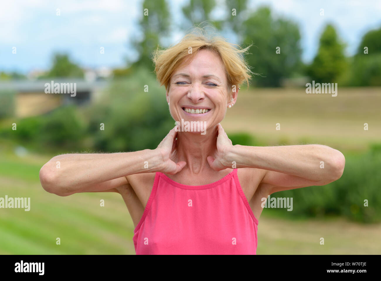 Gerne sportliche Frau, die sich mit den Händen an den Hals, geschlossenen Augen und einem strahlenden Lächeln der Wertschätzung im Freien in einer ländlichen Landschaft Stockfoto