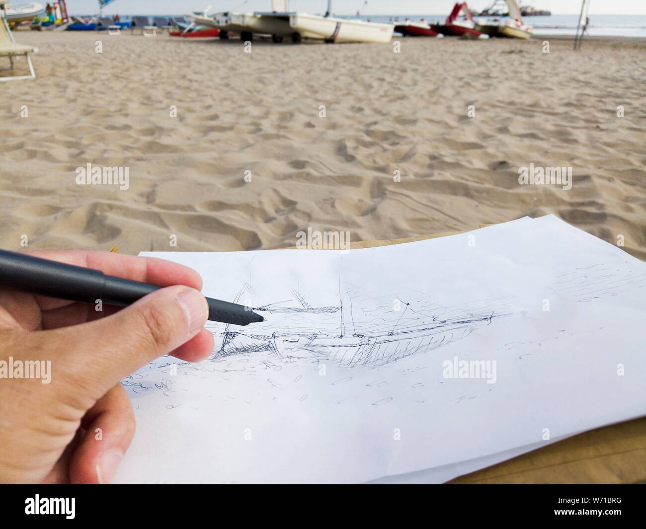 Ein Künstler Hand skizzieren mit einem Stift auf das Papier beim Sitzen am Strand, Außenaufnahme Stockfoto