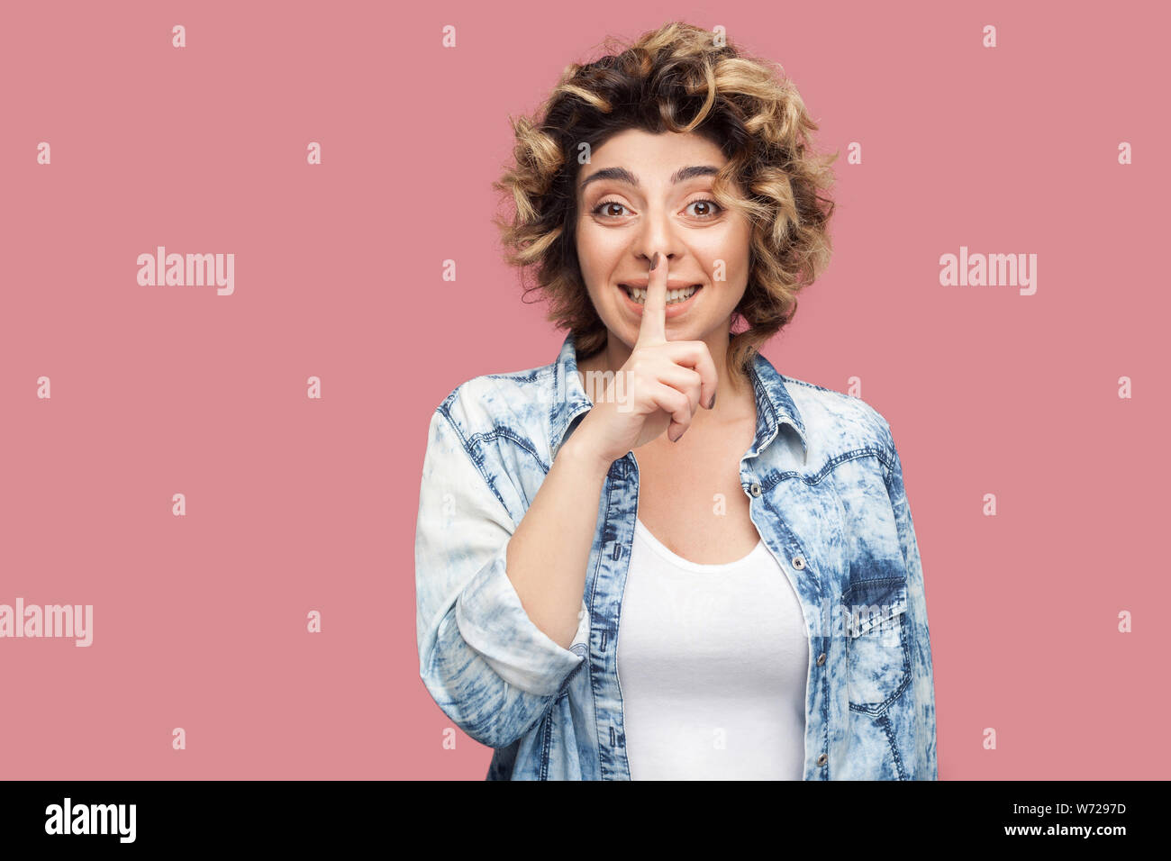 Portrait von sorgen junge Frau mit Curly Frisur im casual blaues Hemd stehen, geheim, und wenn man die Kamera mit dem Finger auf den Lippen geheime Zeichen Stockfoto