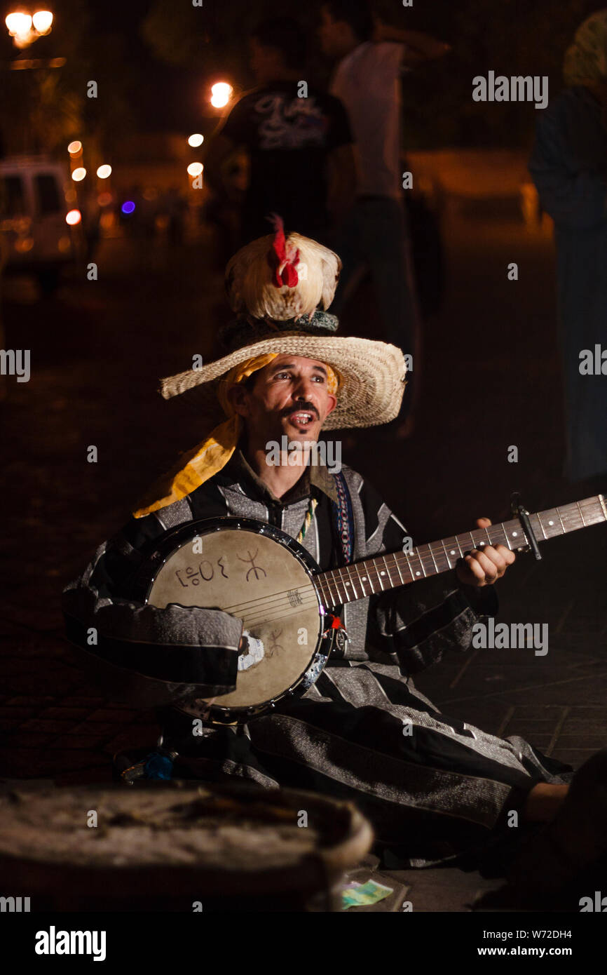 Marokko, Marrakesch. Ein Mann mit einem Hahn auf den Kopf spielt Banjo und singt in Djemaa el Fna Square bei Nacht Stockfoto