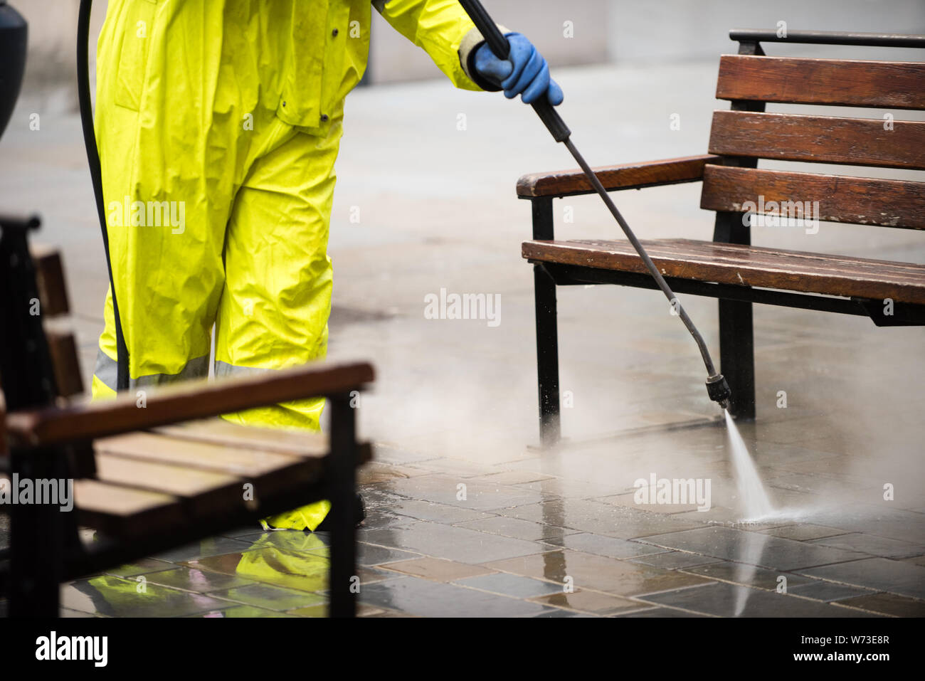 Bürgersteig und Bänke in einer Einkaufsstraße, Jet gewaschen und von einem Arbeiter Gelb hohe vis Kleidung sprühen Wasser aus einem Schlauch gereinigt Stockfoto