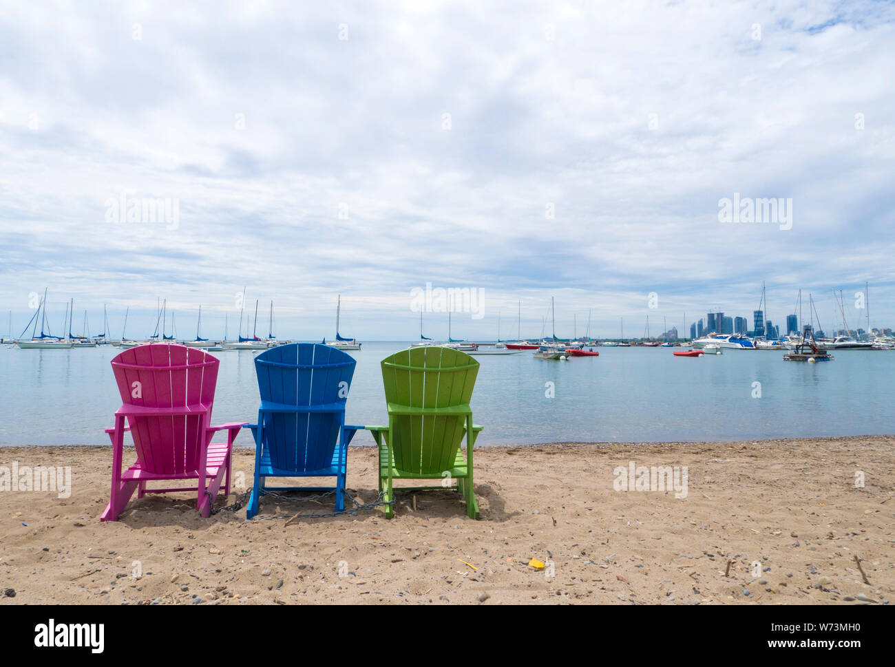 Mehrfarbige Muskoka Cottage Stühle auf dem Sand den Strand, den Hafen und die Innenstadt von Etobicoke im Hintergrund suchen Stockfoto