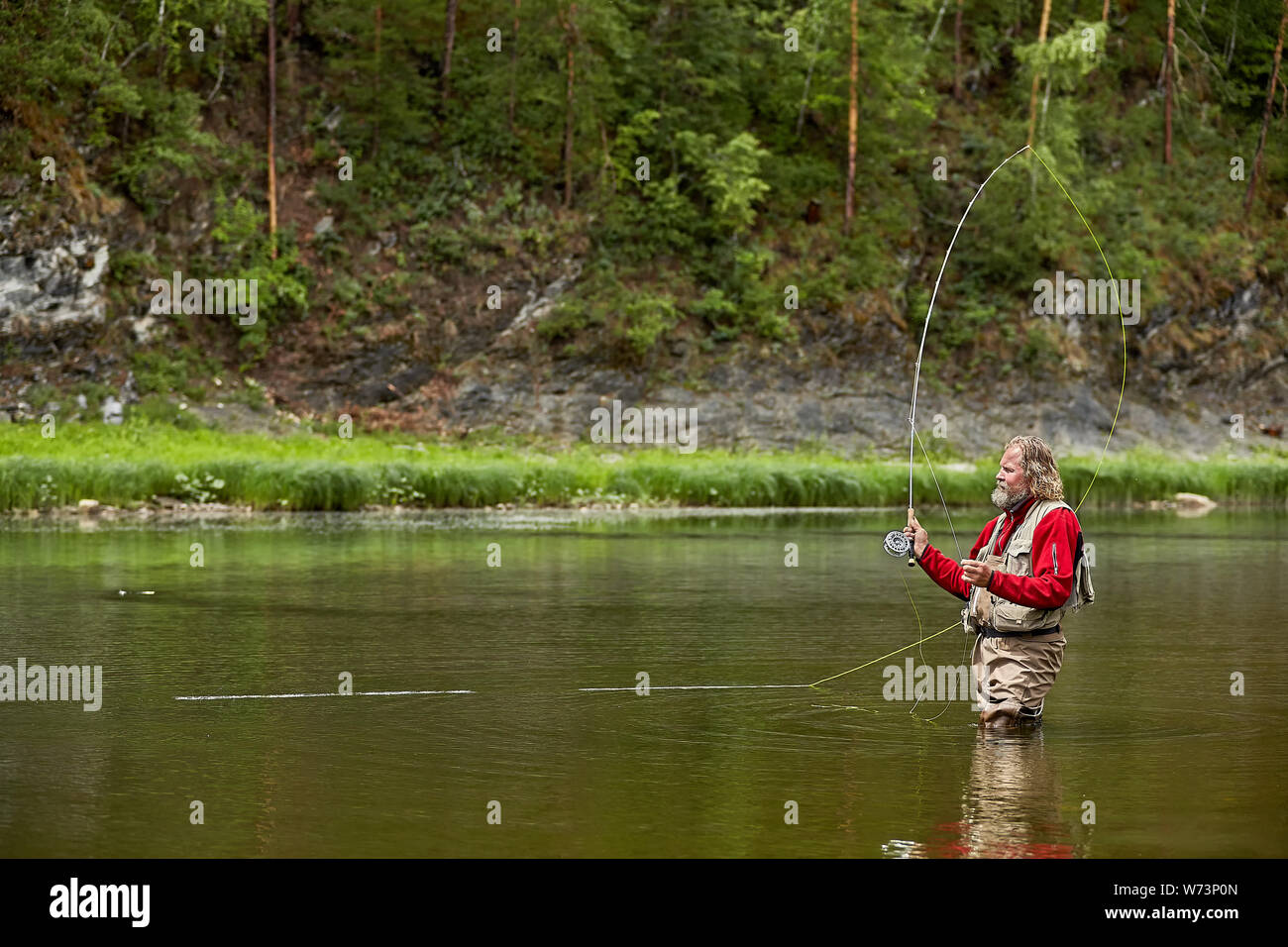Reife bärtige Mann in Form von wasserdichter Kleidung im Stehen im Fluss im Wald beim Fliegenfischen, eco - Tourismus. Stockfoto