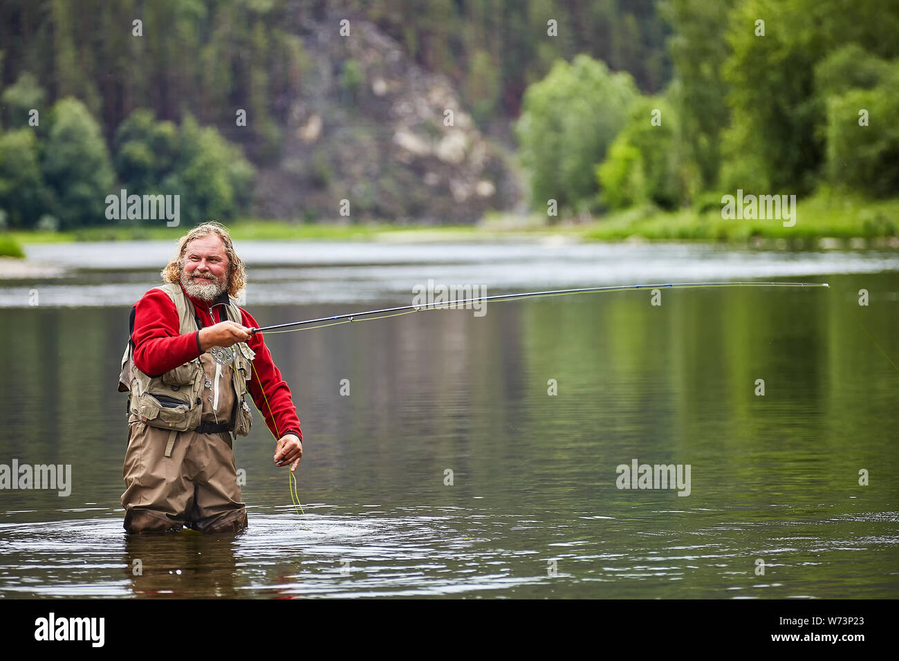 Whire nass Reife bärtige Mann steht im Wasser in wasserdichte Kleidung und Vorbereitung Fliegenfischen anzufangen, er lächelt, eco Tourismus. Stockfoto