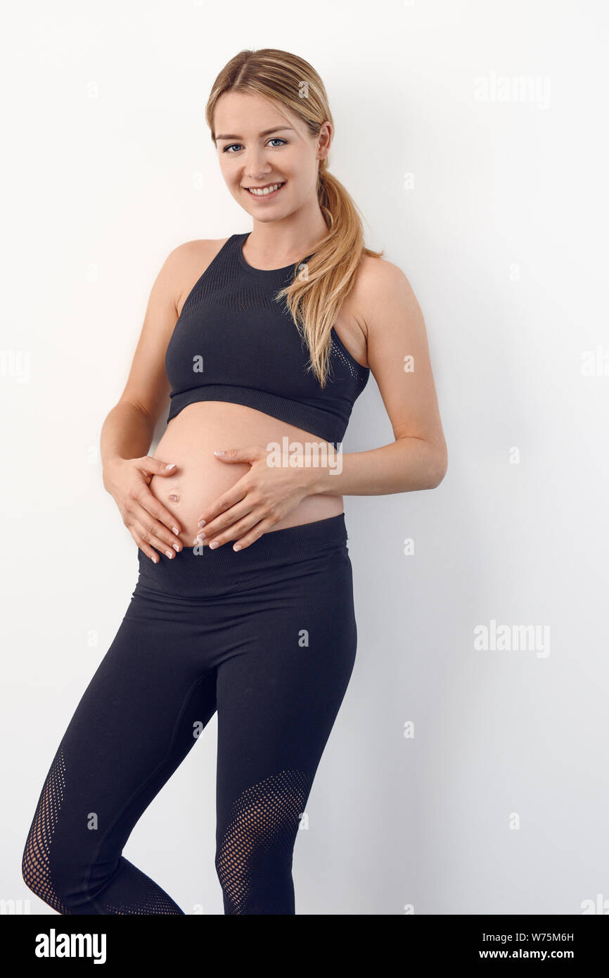 Glücklichen gesunden jungen schwangeren Frau in Schwarz Sportbekleidung auf den Hüften in die Kamera schaut mit einem strahlenden Lächeln Stockfoto